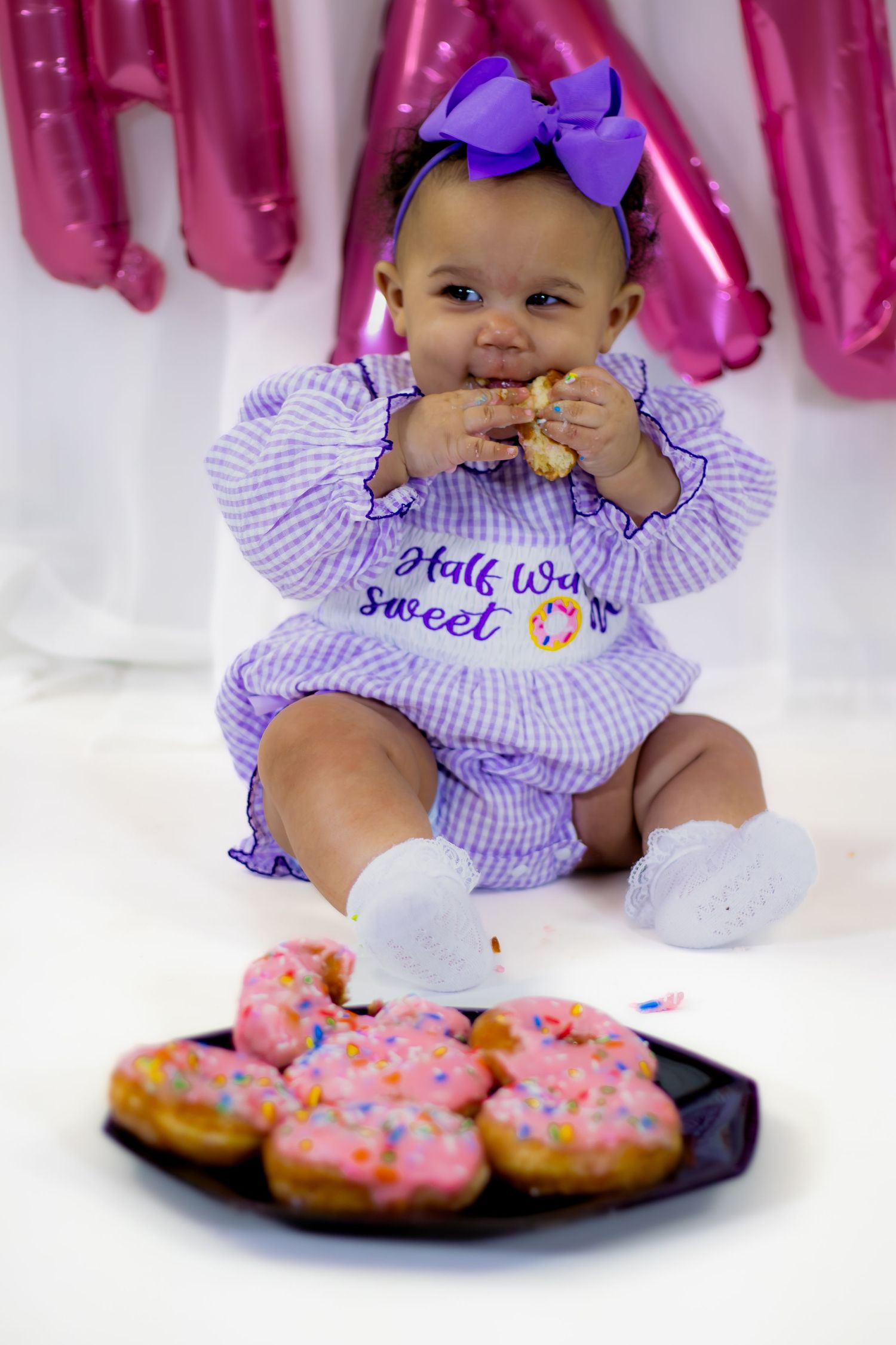 Baby girl in Half Way to Sweet One romper munches pink sprinkle donut at party.