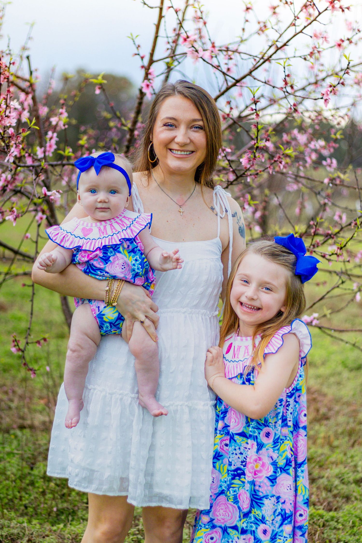 Smiling mother in white dress holding infant beside older daughter in blue floral dress near pink blossoms.