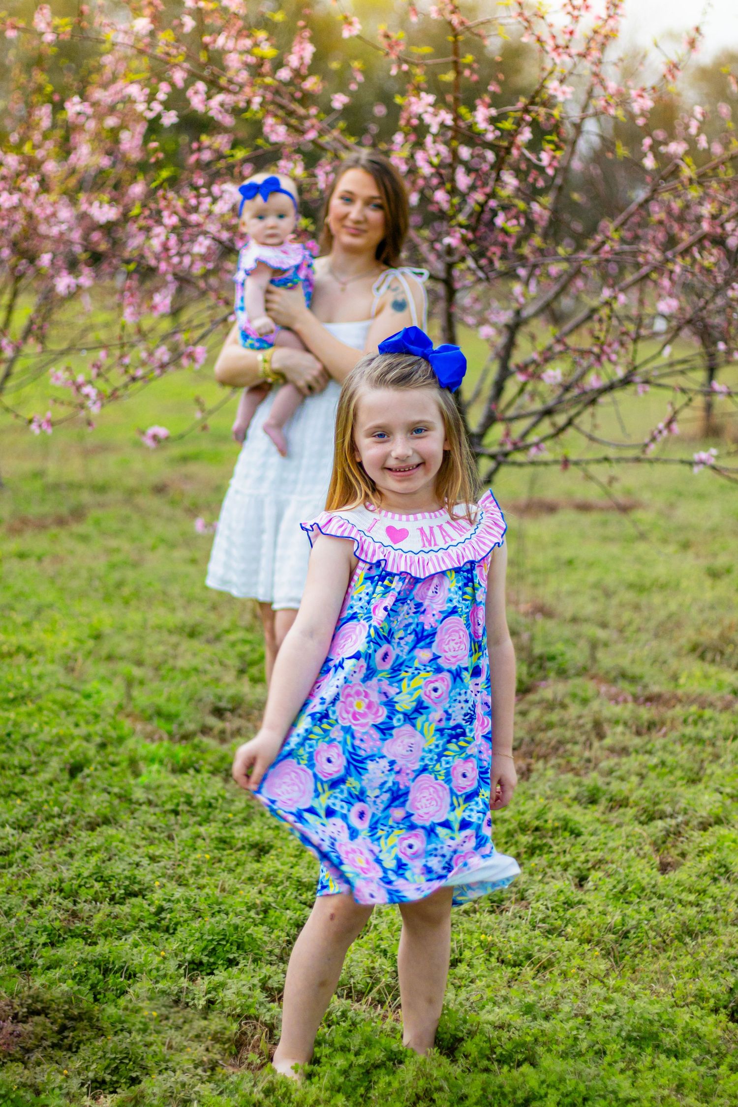 Young girl in blue floral dress smiles as mother holds baby sister near pink flowering trees.