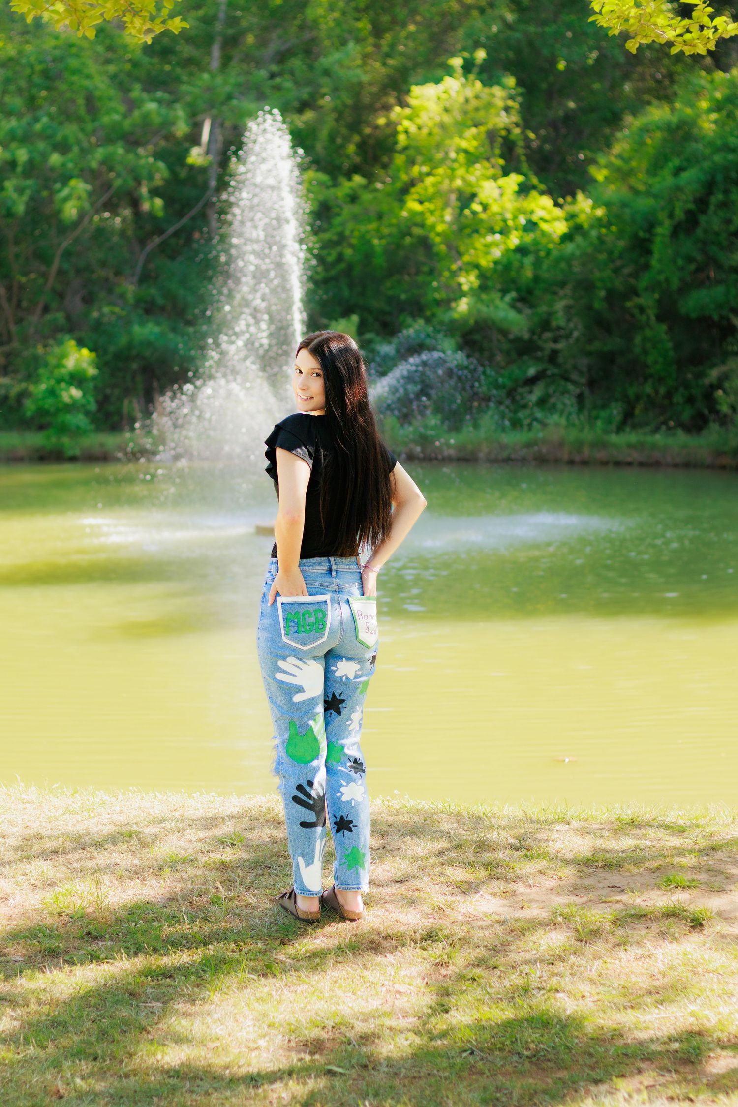Young woman in painted jeans stands by a park fountain, surrounded by lush green trees on a sunny day.