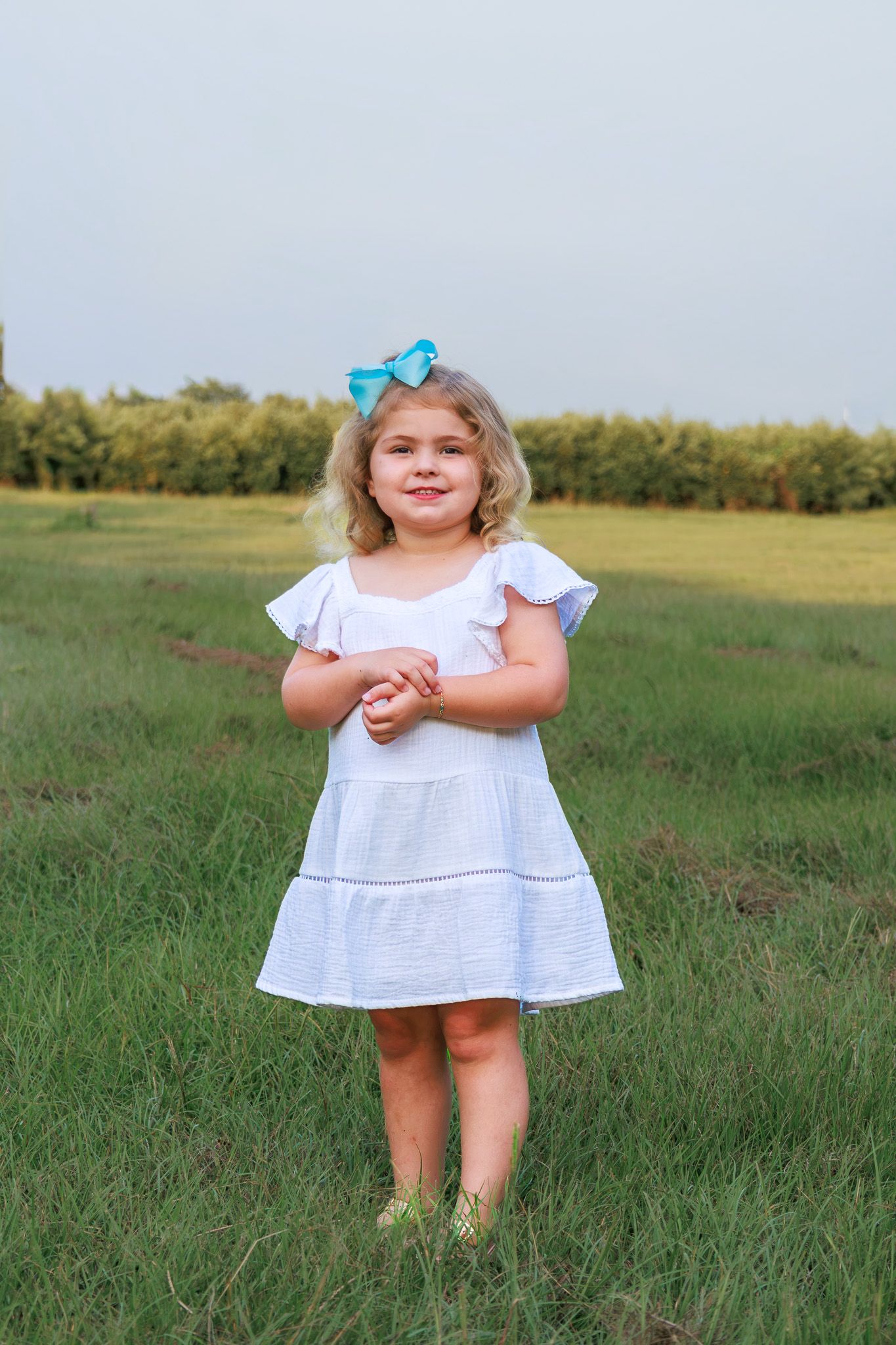 Young girl in white dress with blue bow stands smiling in green field.