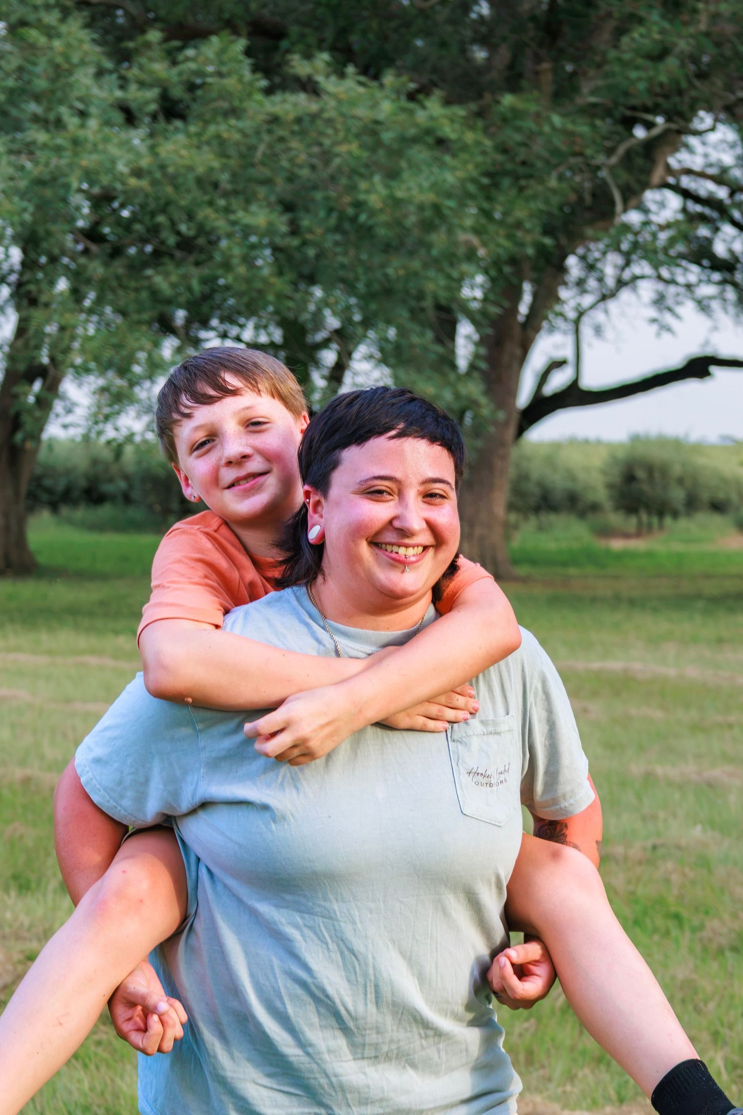 A woman gives a smiling boy a piggyback ride outdoors in a green field with trees in the background.