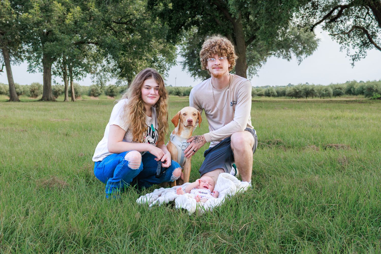Two teens and a dog pose with a newborn baby in a lush green field surrounded by trees.