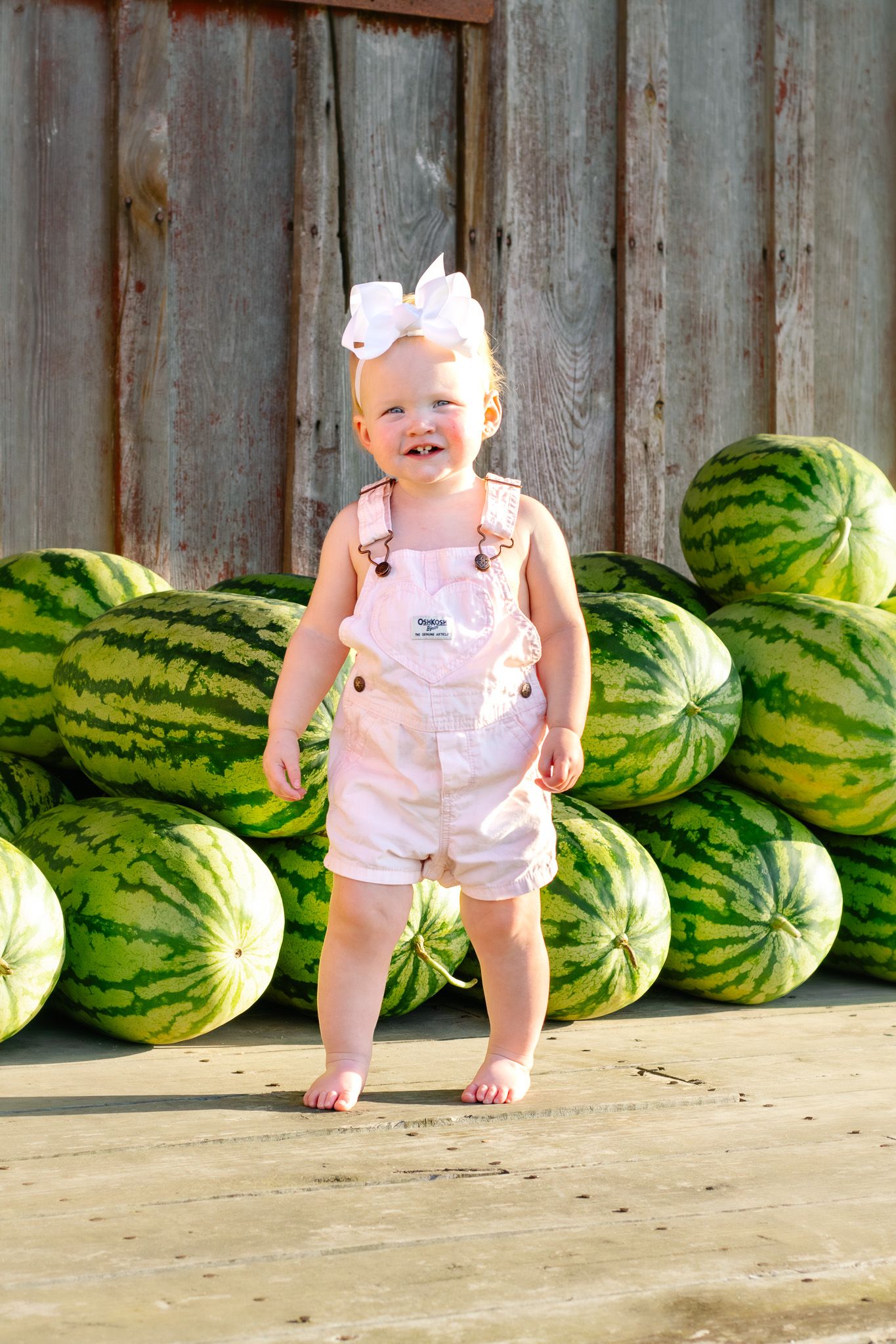 Smiling toddler girl in pink overalls and white bow stands barefoot in front of stacked watermelons by a wooden barn.