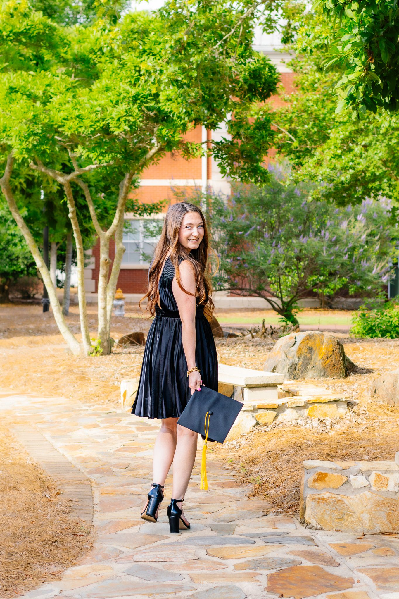 Smiling graduate in black dress holding cap with gold tassel on stone pathway surrounded by trees.