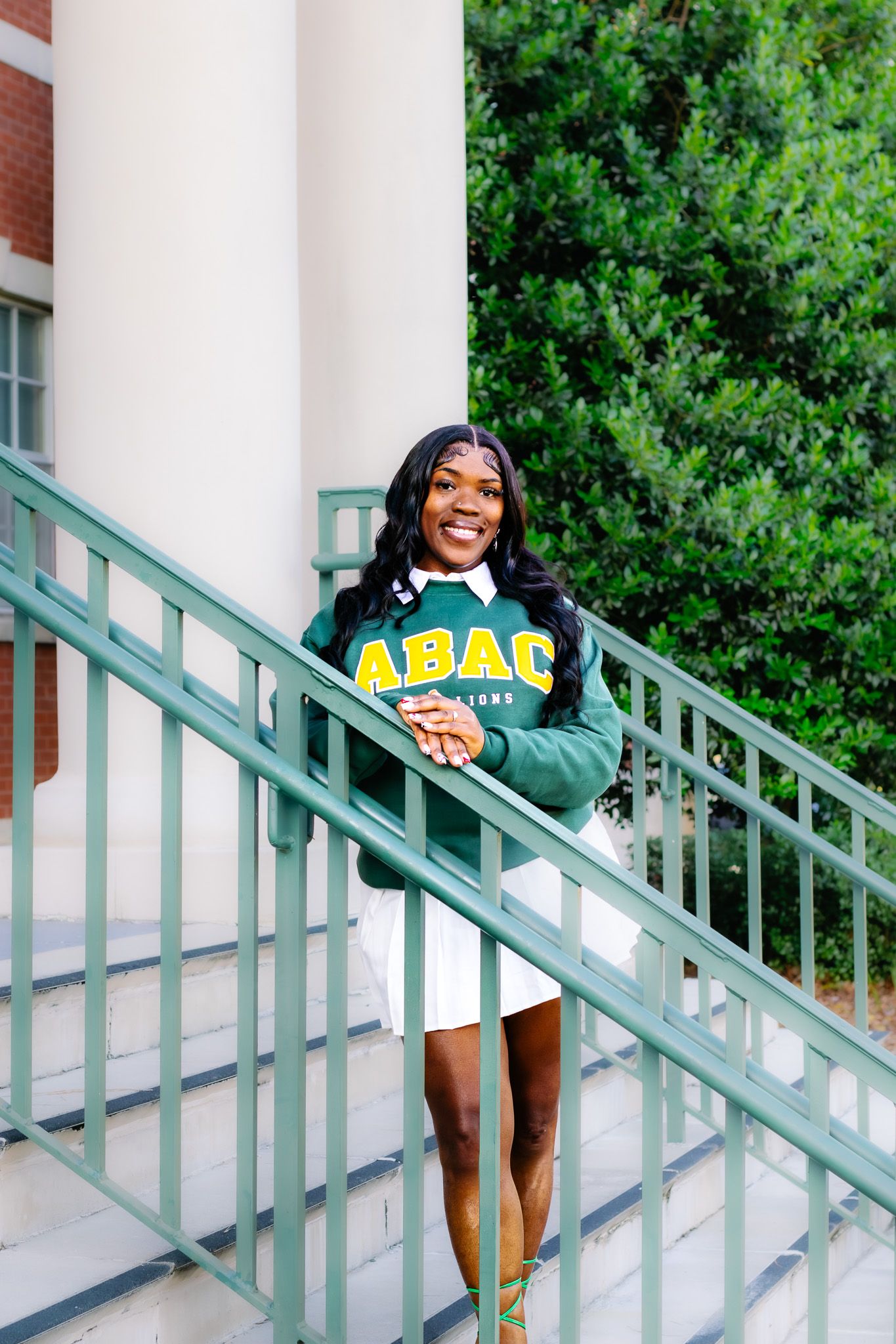 Smiling student wearing an ABAC green and yellow college sweatshirt poses on green staircase railing outdoors.
