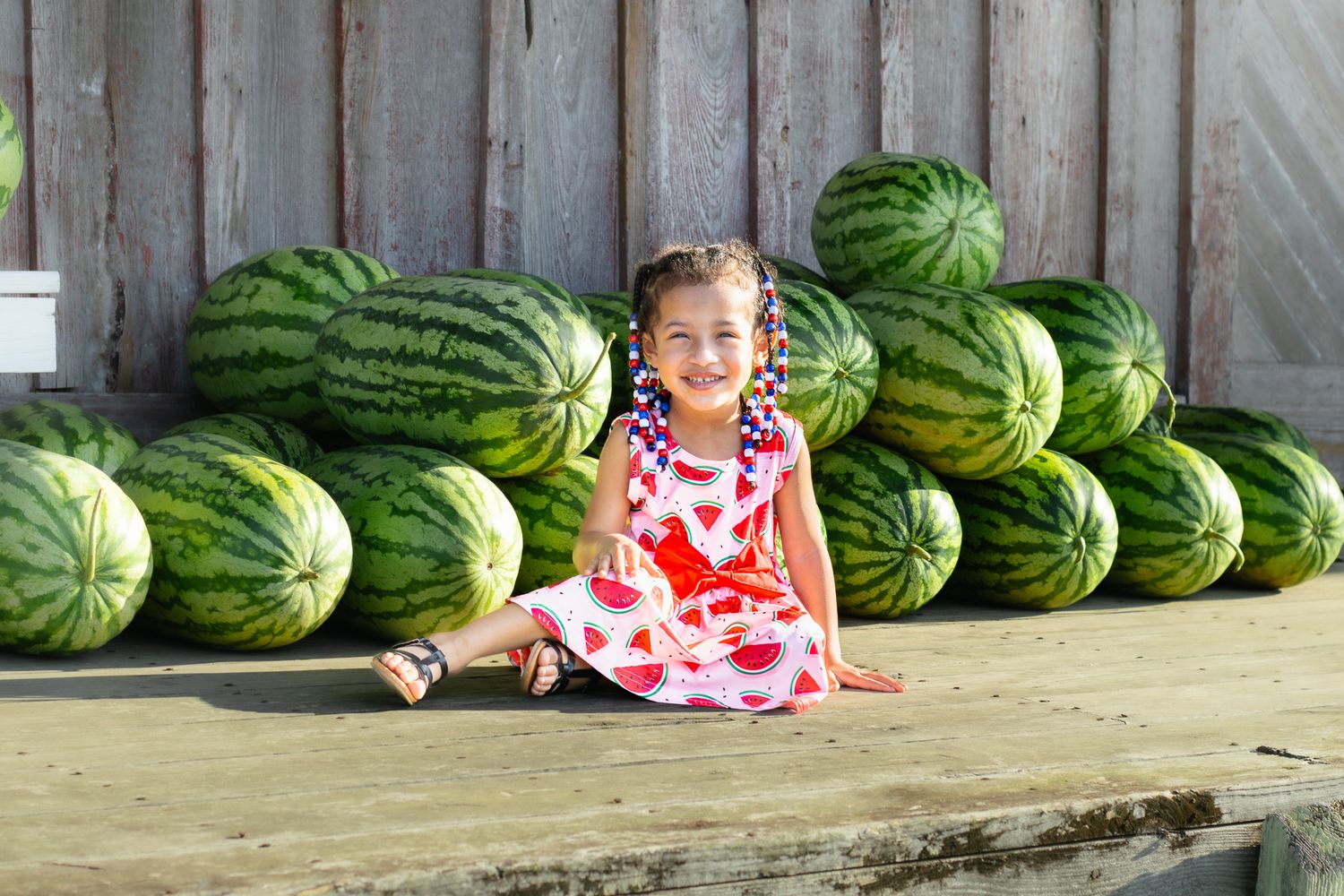 Young girl in floral dress sits smiling among large stacked watermelons at a farm stand.