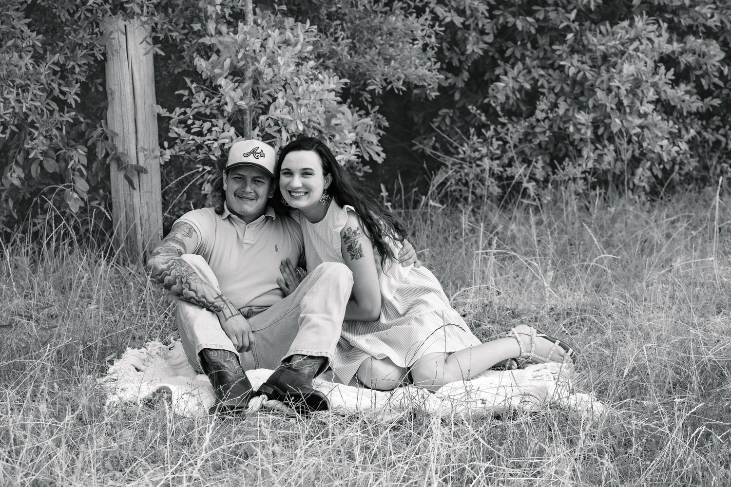 Black and white photo of a couple sitting together on a blanket in a grassy field near a wooden post.