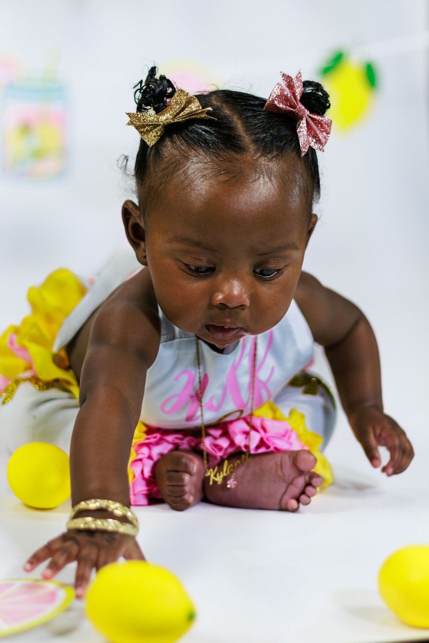 Baby girl in lemon themed birthday outfit crawling toward yellow lemons on white floor.