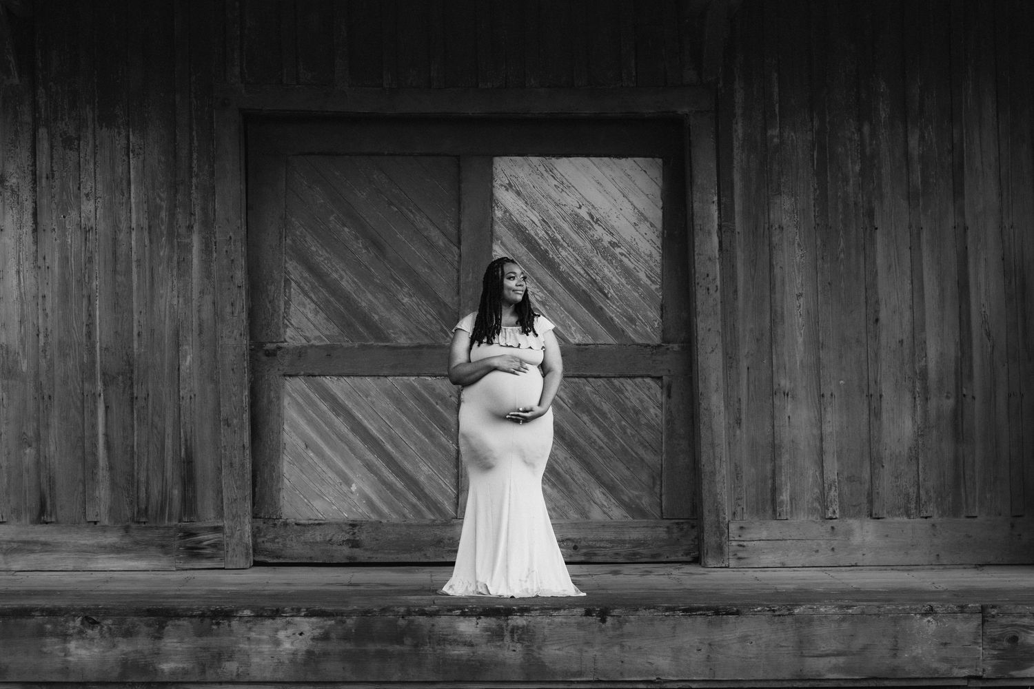 Pregnant woman in white gown poses by rustic wooden barn doors in elegant black and white maternity photo.