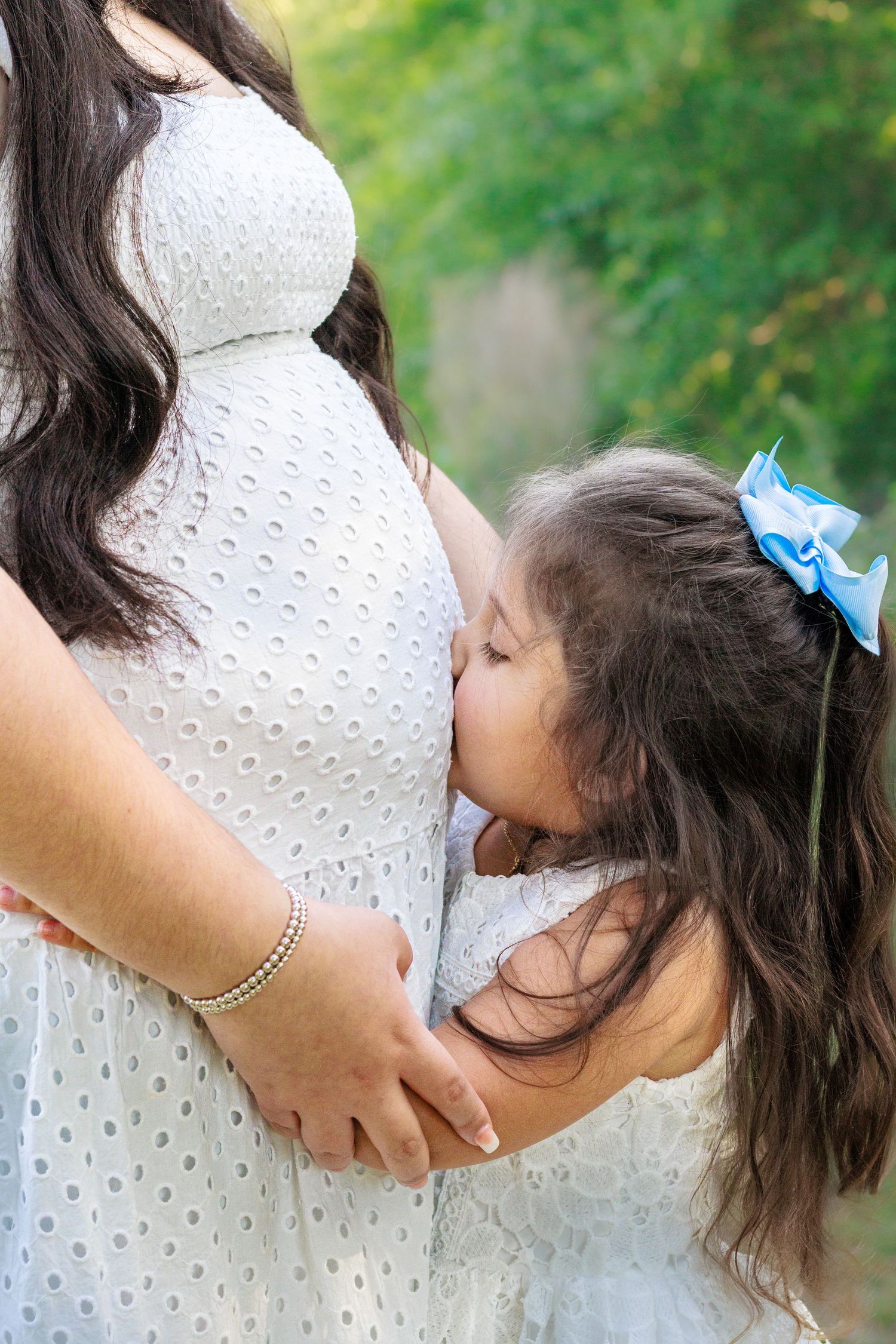 A young girl in white dress kisses her pregnant mothers baby bump outdoors during a maternity photo session.