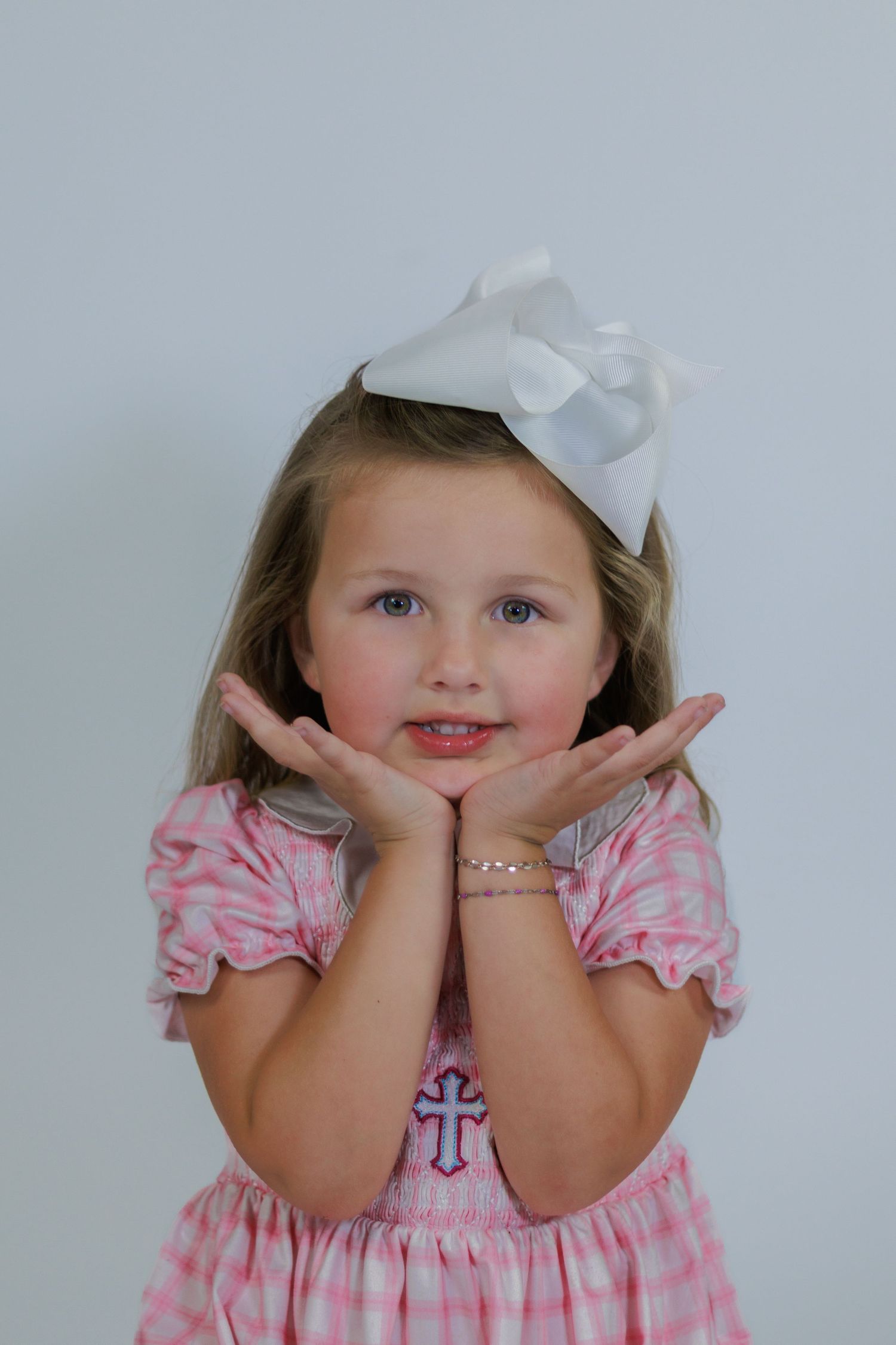 Young girl in pink gingham dress with white bow poses with hands under chin, wearing a cross bracelet.