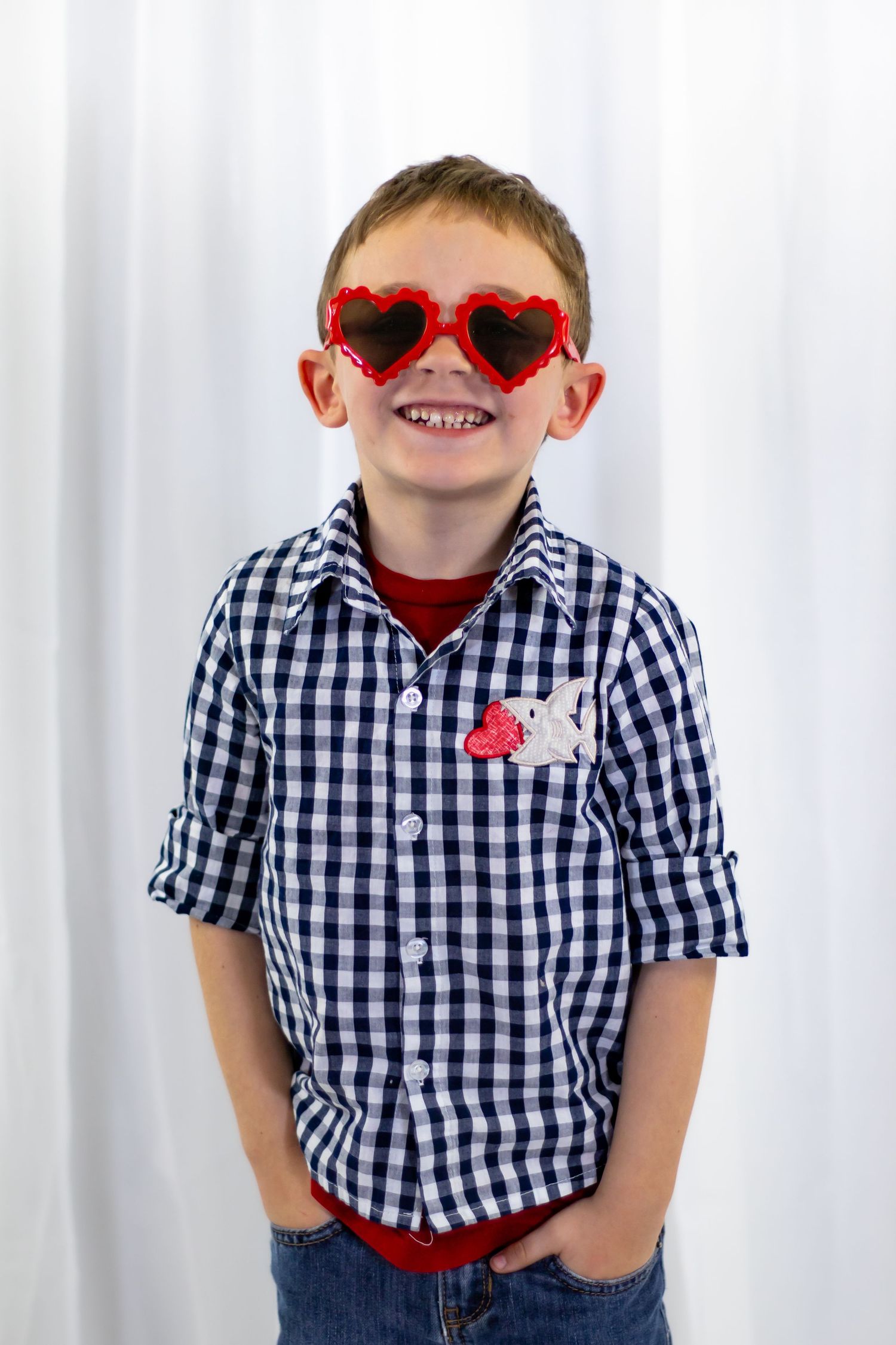 Smiling boy in gingham shirt wears red heart-shaped sunglasses with hands in pockets on white backdrop.