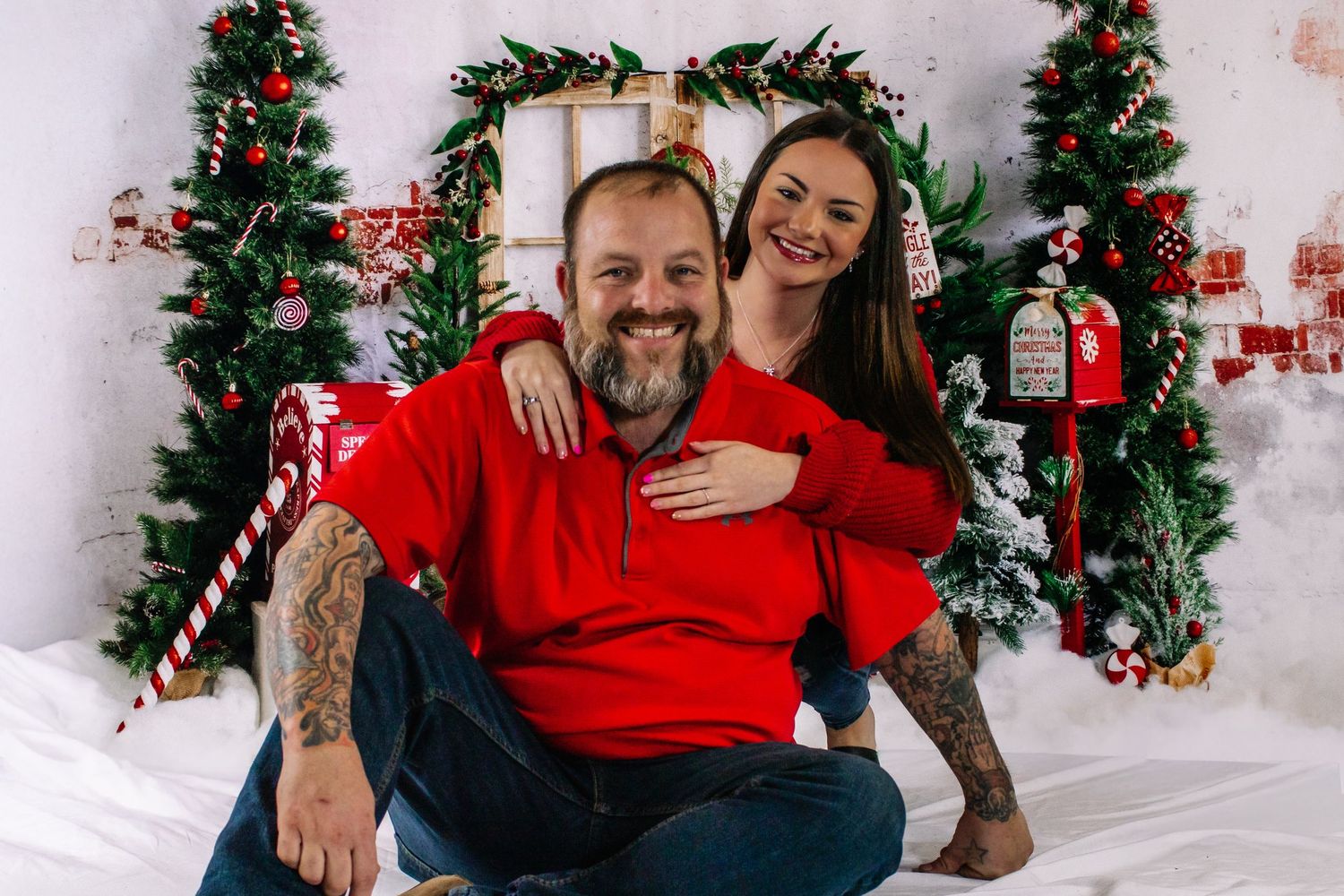 Tattooed couple in red shirts smiling together in front of Christmas holiday photo backdrop.