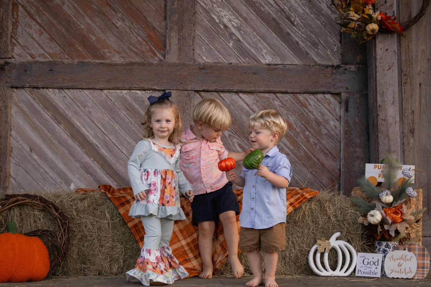 Three young children stand together on hay bales with colorful fall pumpkin patch decorations.