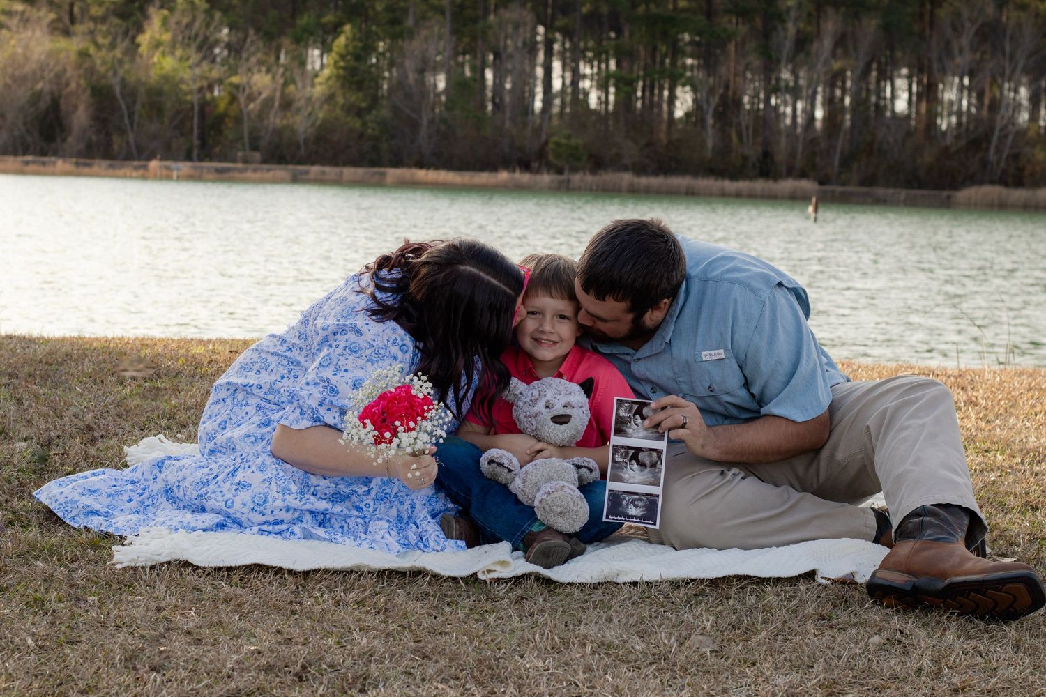 Family of three sharing tender moment on blanket by lake, parents kissing child holding ultrasound photos.