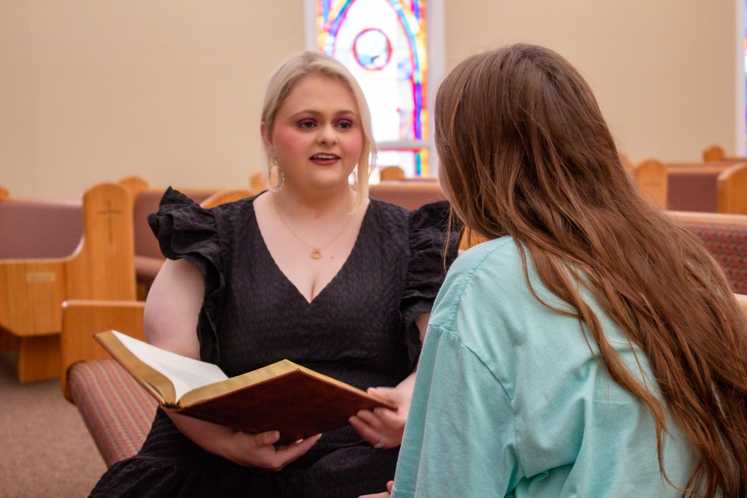 Two women sit in church pews reading a Bible together near a vibrant stained glass window.