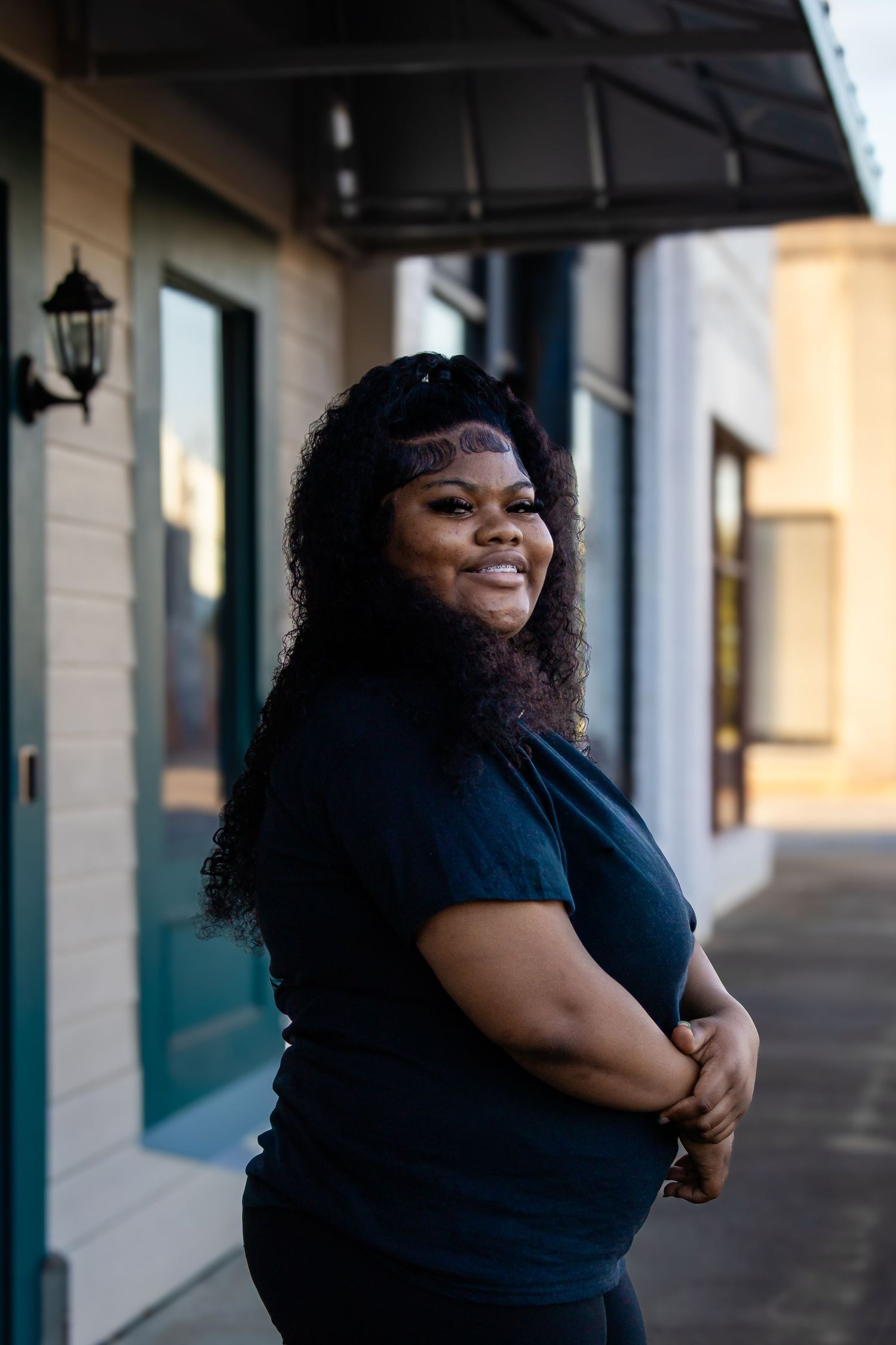 Smiling young Black woman with headband in navy top posing on urban sidewalk outside teal door.