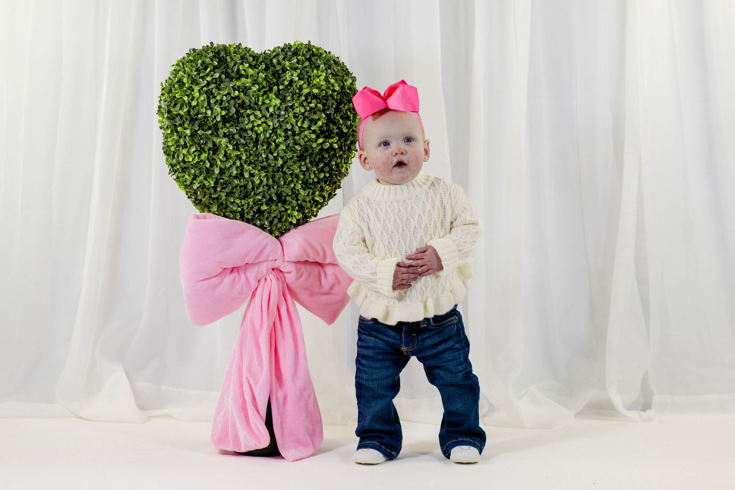 Curious baby girl in white sweater and pink bow standing next to a heart-shaped topiary with pink bow.