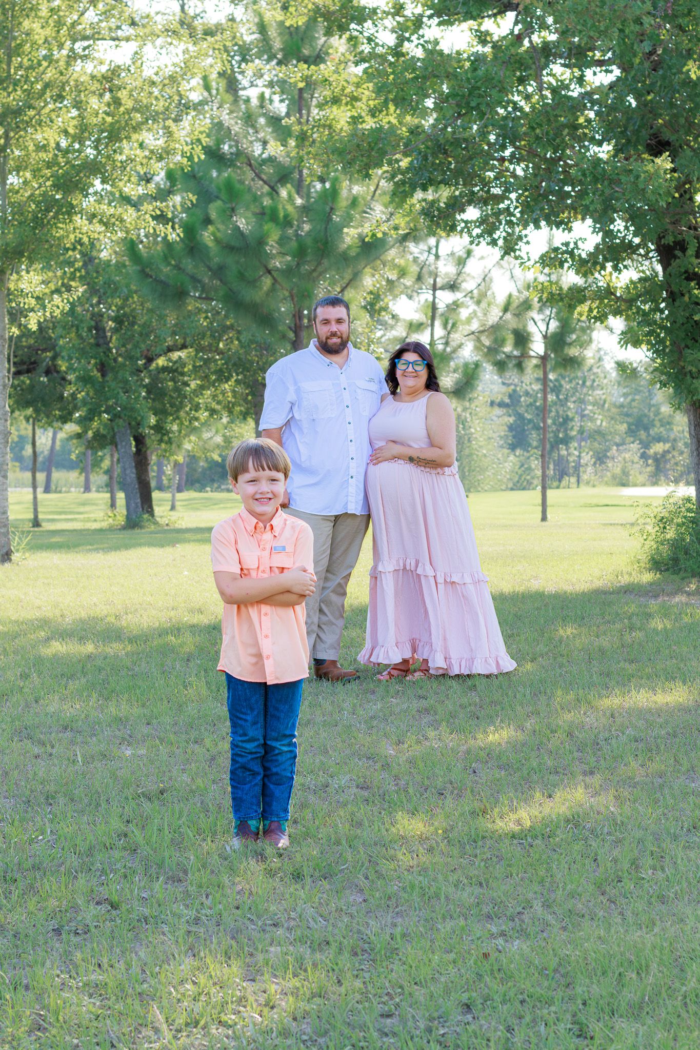 Young boy stands confidently in front of his parents during a family maternity photo session in a park.