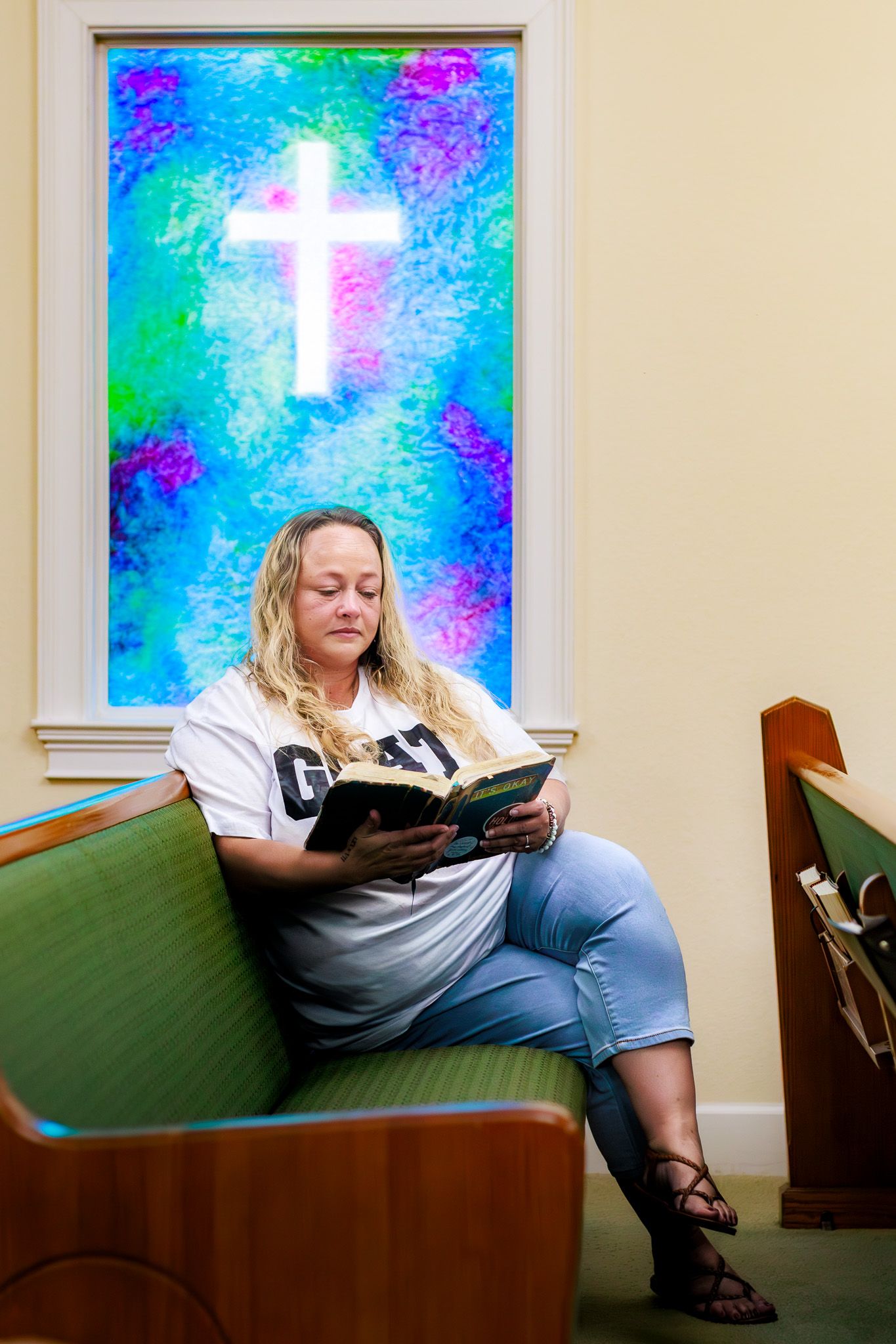 Woman reading Bible in church pew with colorful stained glass cross window behind her.