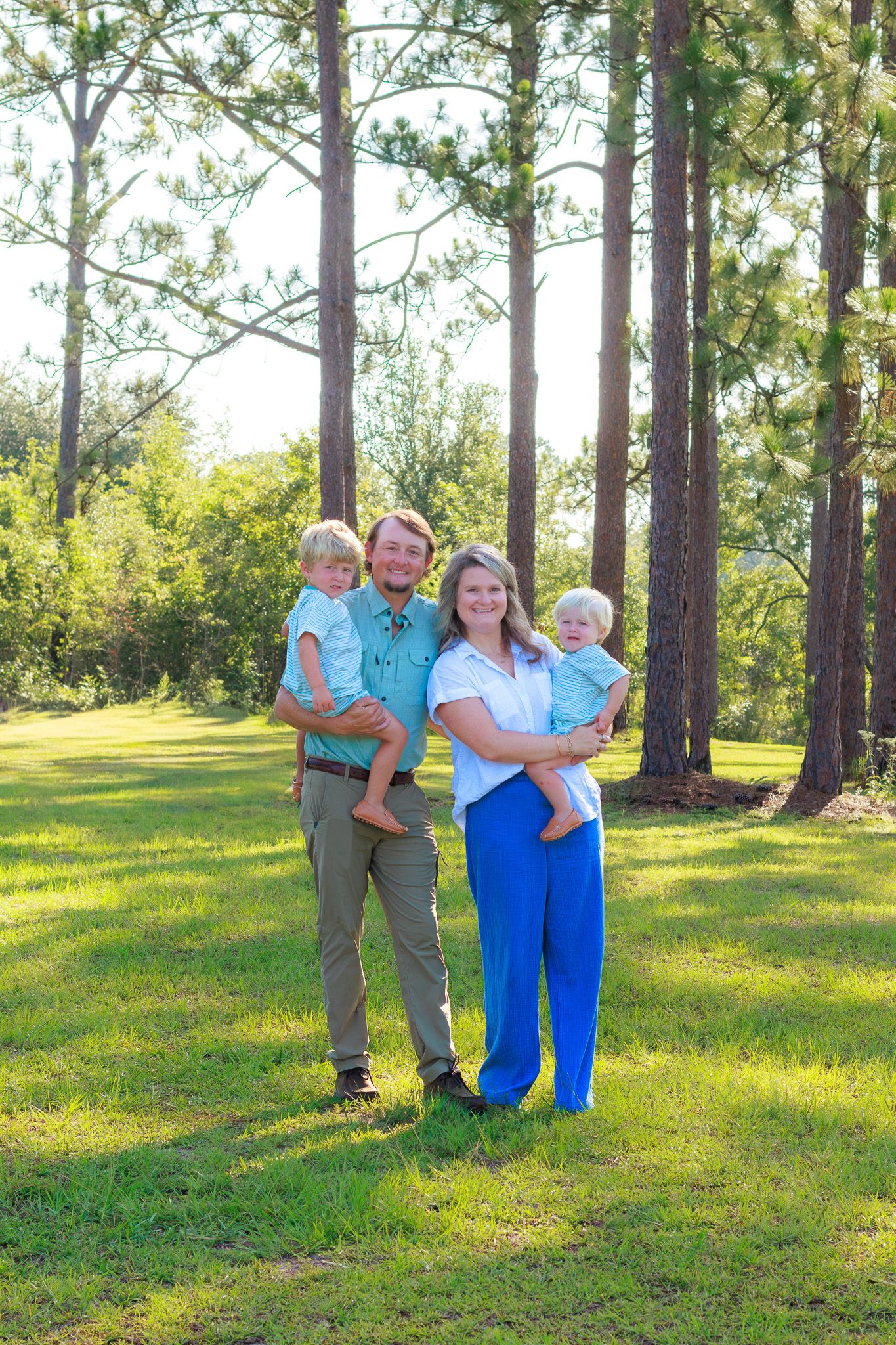 Family of four poses outdoors among tall pine trees on green grass, parents holding two young children.