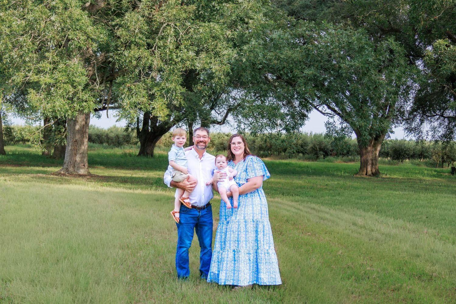 Family of four posing outdoors on green grass with large trees, wearing coordinating blue outfits on a sunny day.