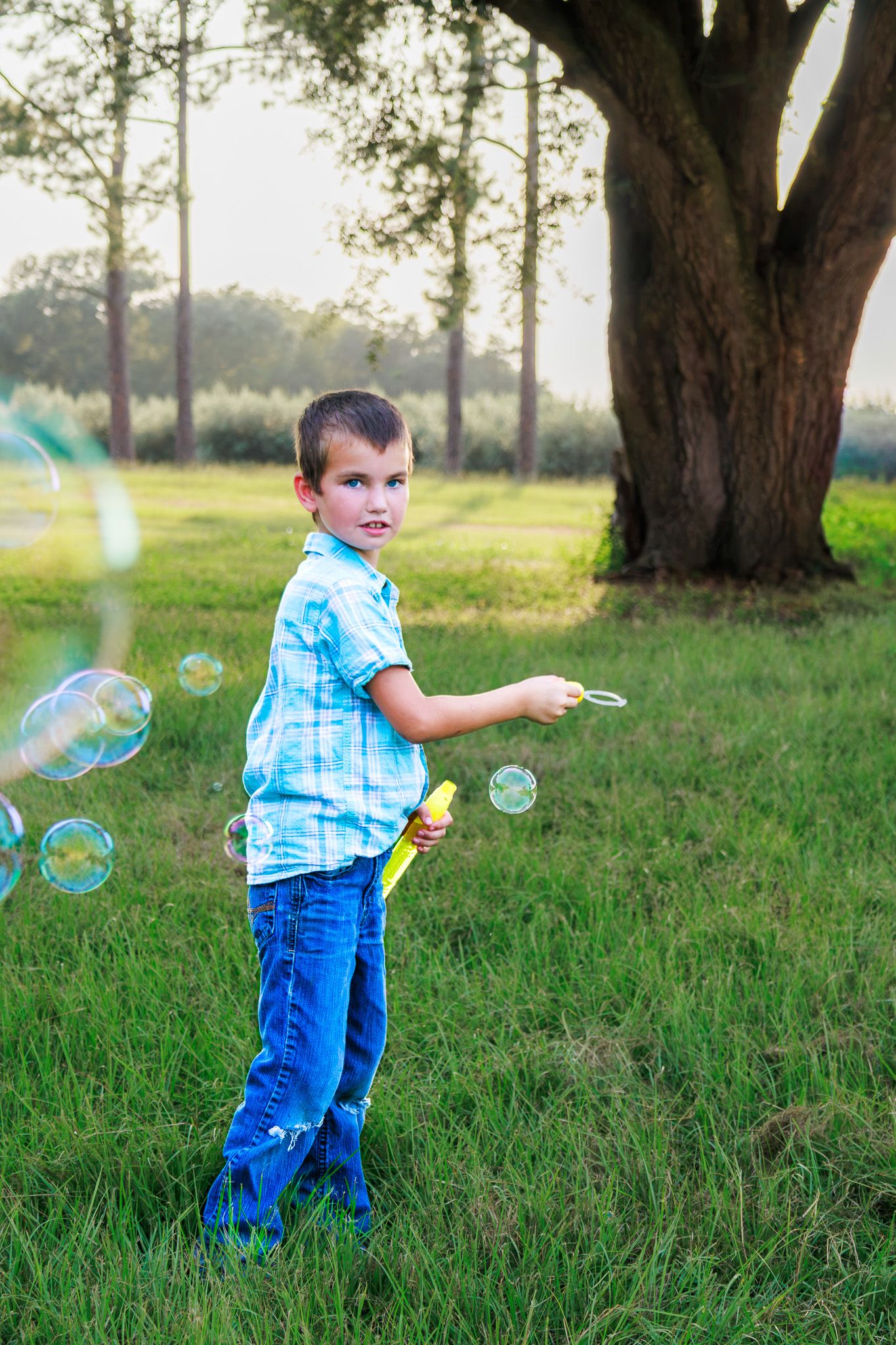 A young boy in a blue shirt and jeans blows bubbles with a toy wand in a sunny green park.
