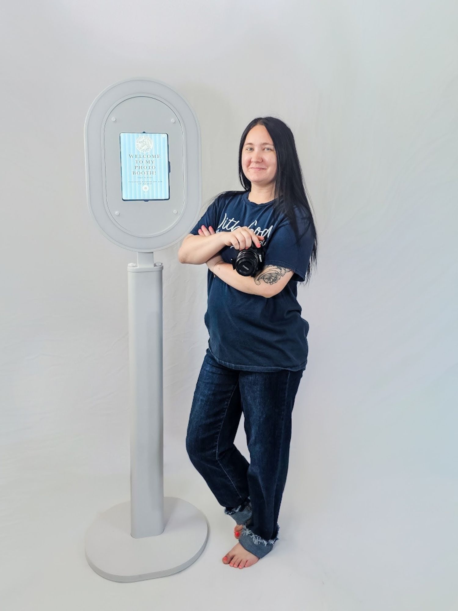 Woman with long dark hair and tattoos stands beside a modern photo booth kiosk on a gray pedestal.