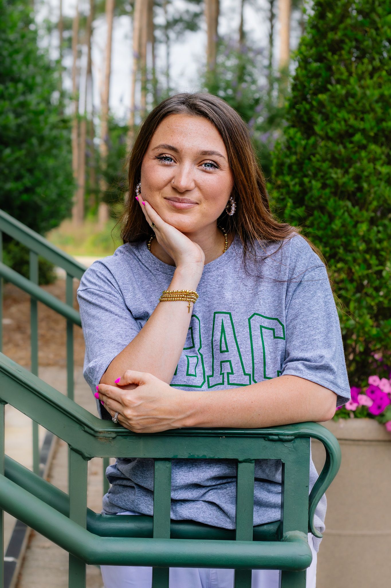 Young woman in gray BAC college t-shirt smiles while leaning on green railing outdoors.