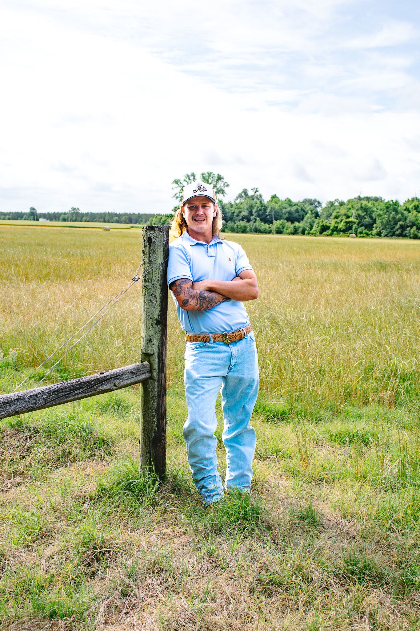 A smiling man in a light blue polo and cap leans against a wooden fence post in a rural field.