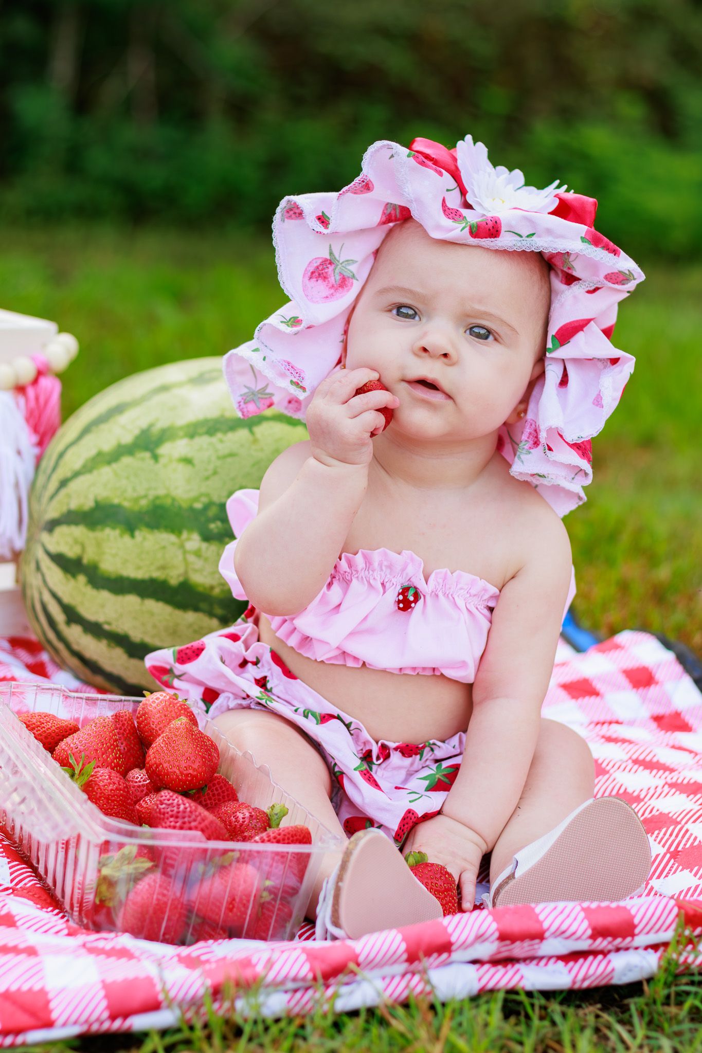 Adorable baby in floral hat tastes a strawberry while sitting on red checkered blanket with watermelon outside.
