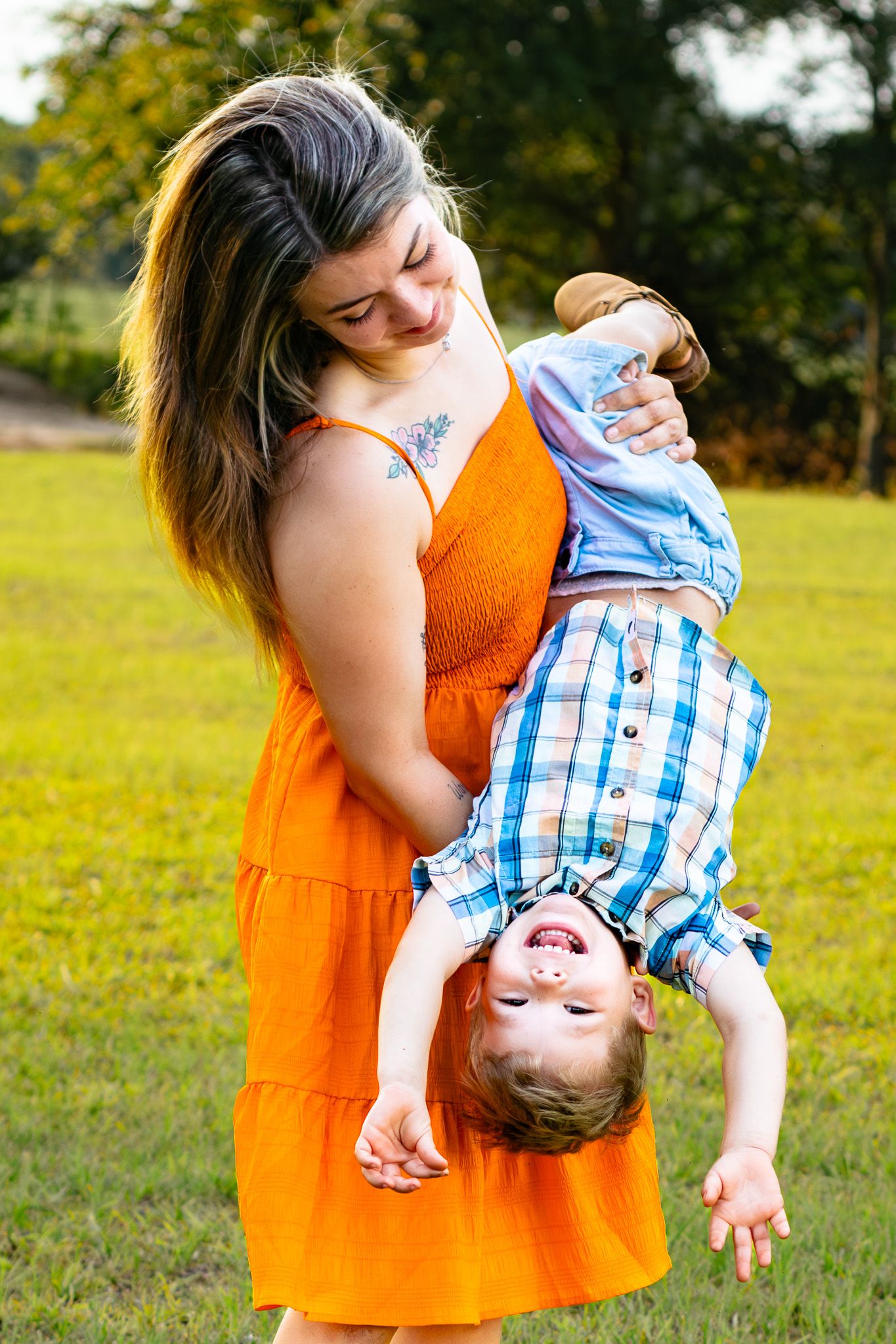 A laughing young boy is held upside down by a smiling woman in an orange dress in a sunny green park.