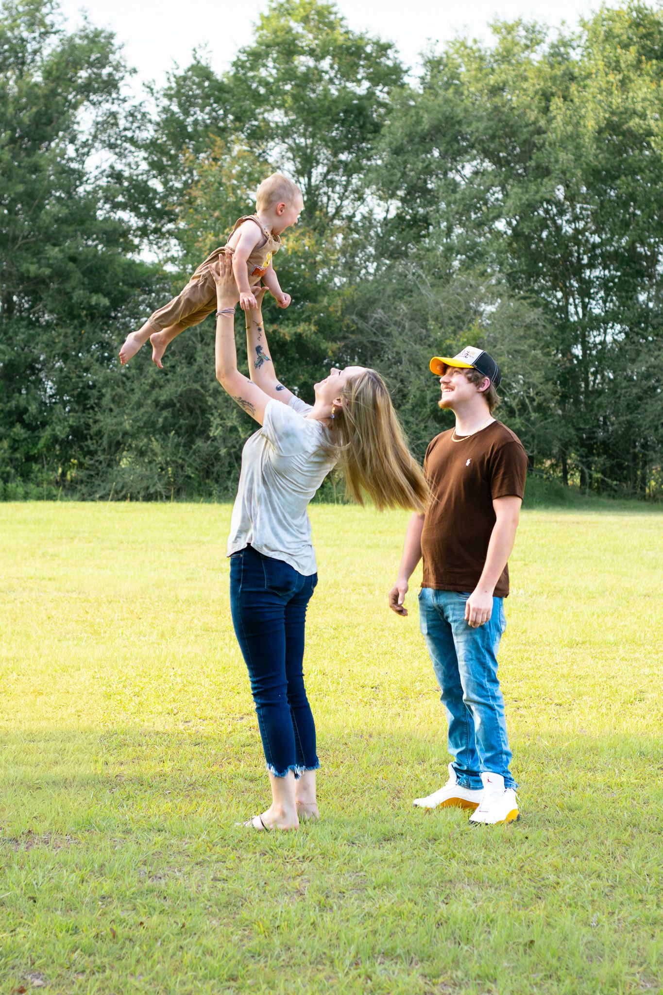 A young family plays outdoors on green grass, with a woman lifting a baby in the air while a man watches nearby.