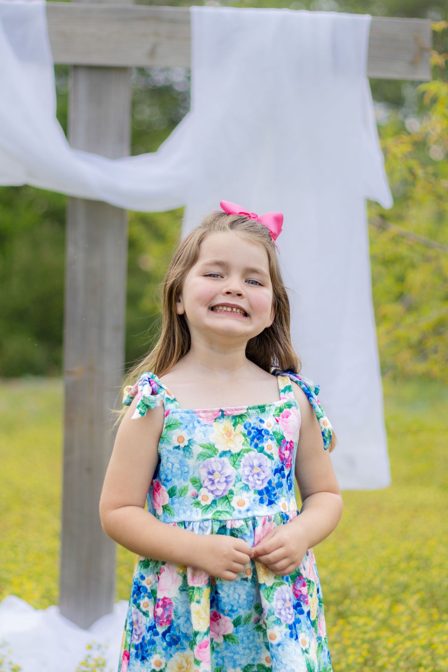 Happy young girl with pink bow wearing blue floral dress smiling outdoors near a white-draped wooden cross.