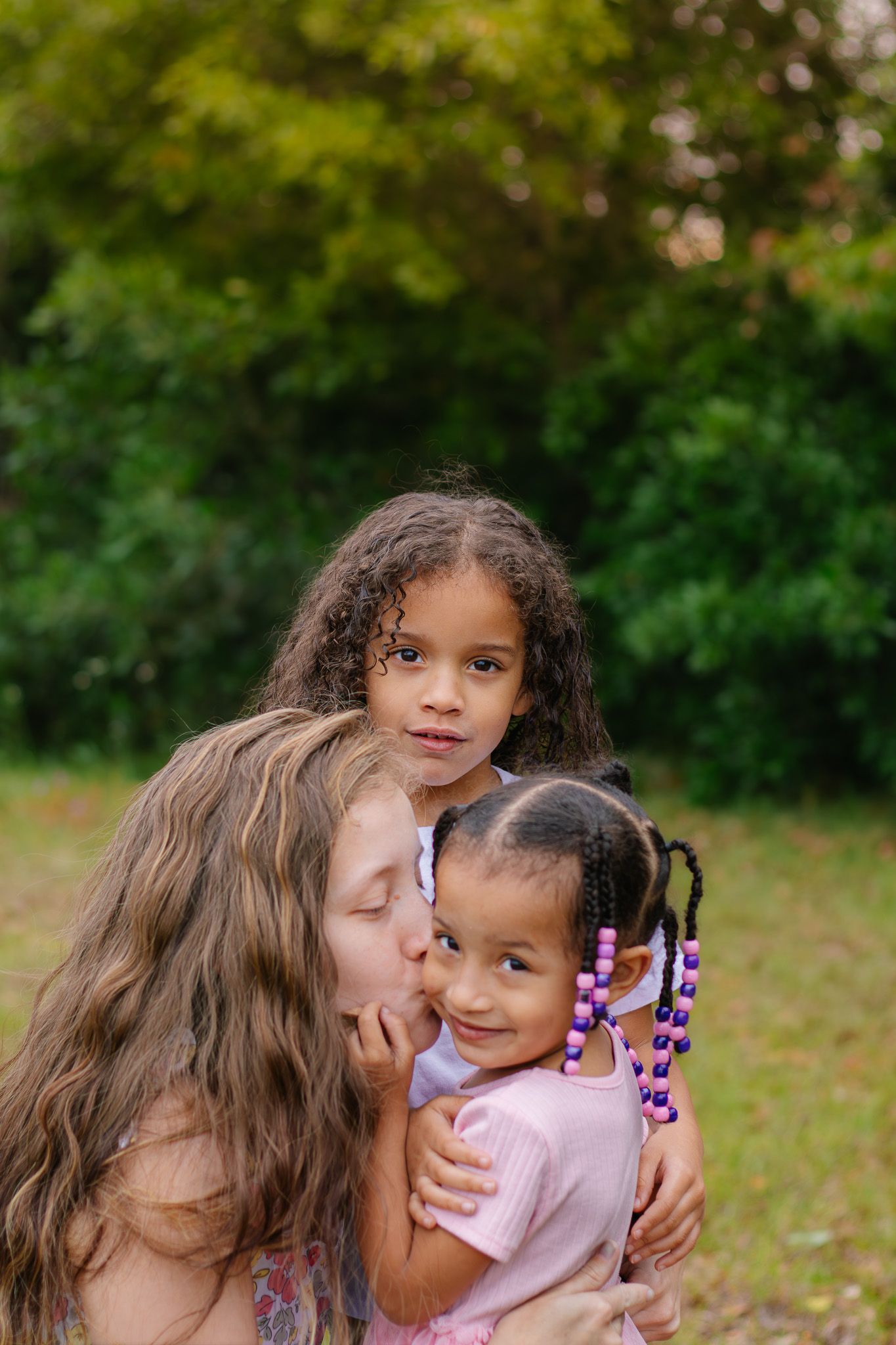 Woman kissing young girl's cheek while hugging two multiracial children outdoors in green park setting.
