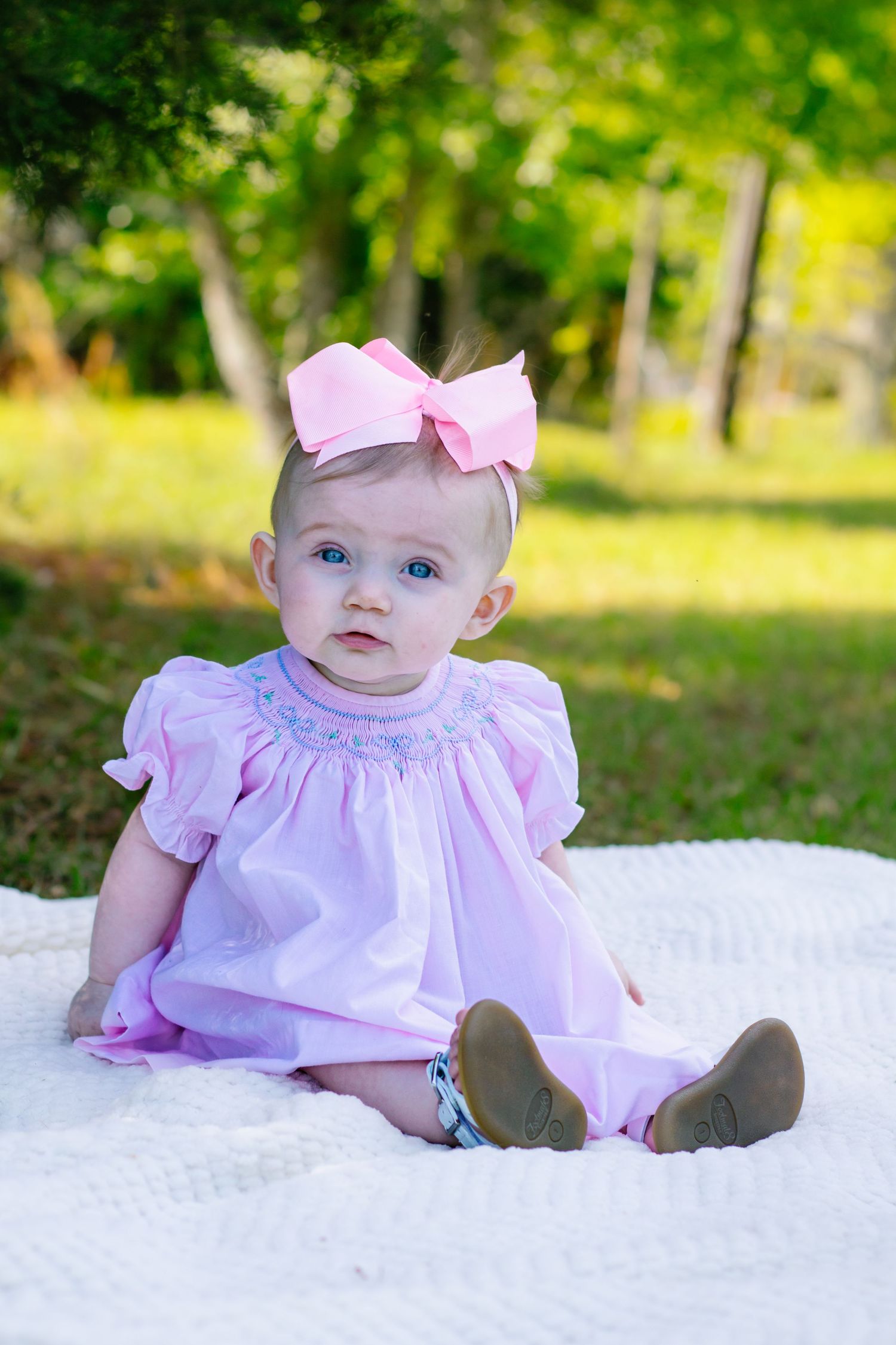Baby girl in pink smocked dress and bow headband sitting on white blanket outdoors in green park.