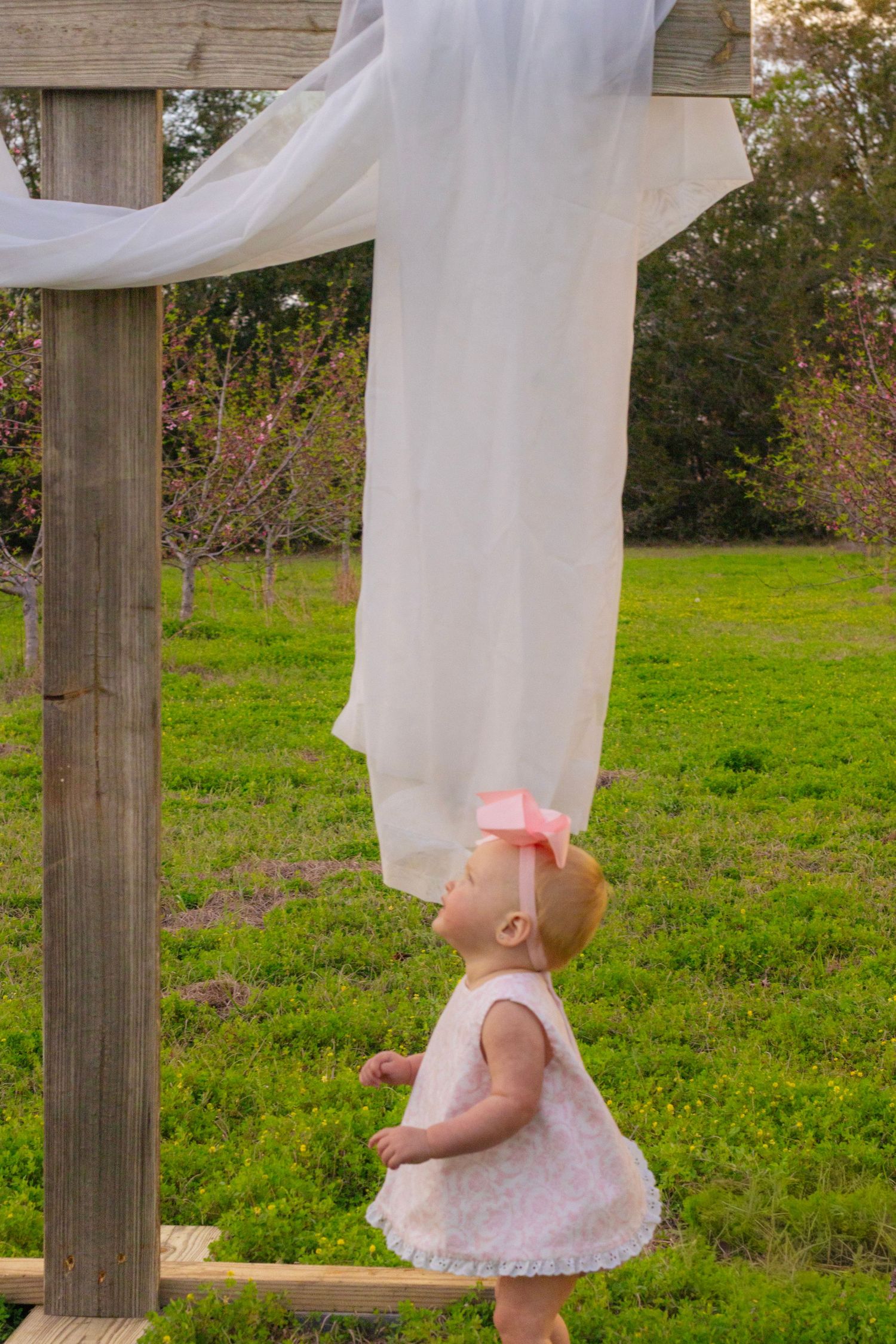 Baby girl in white dress and pink bow looks up at white fabric draped on a large wooden cross.