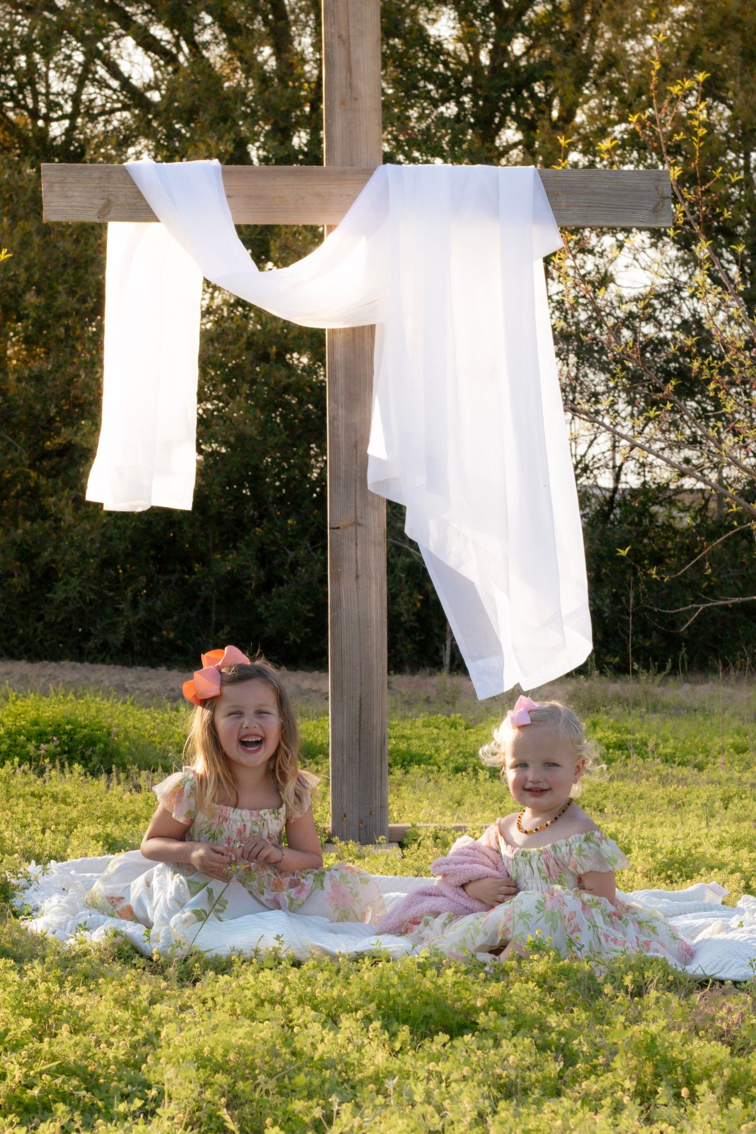 Two laughing girls in matching floral dresses posed beside a draped wooden cross in a sunny field.