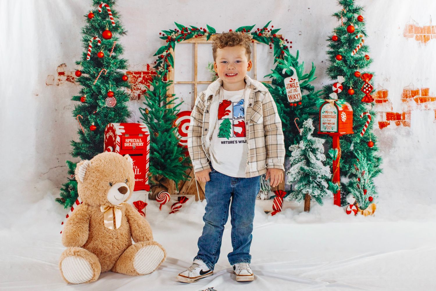 A young boy in a plaid jacket stands next to a large teddy bear in a Christmas-themed photo studio.