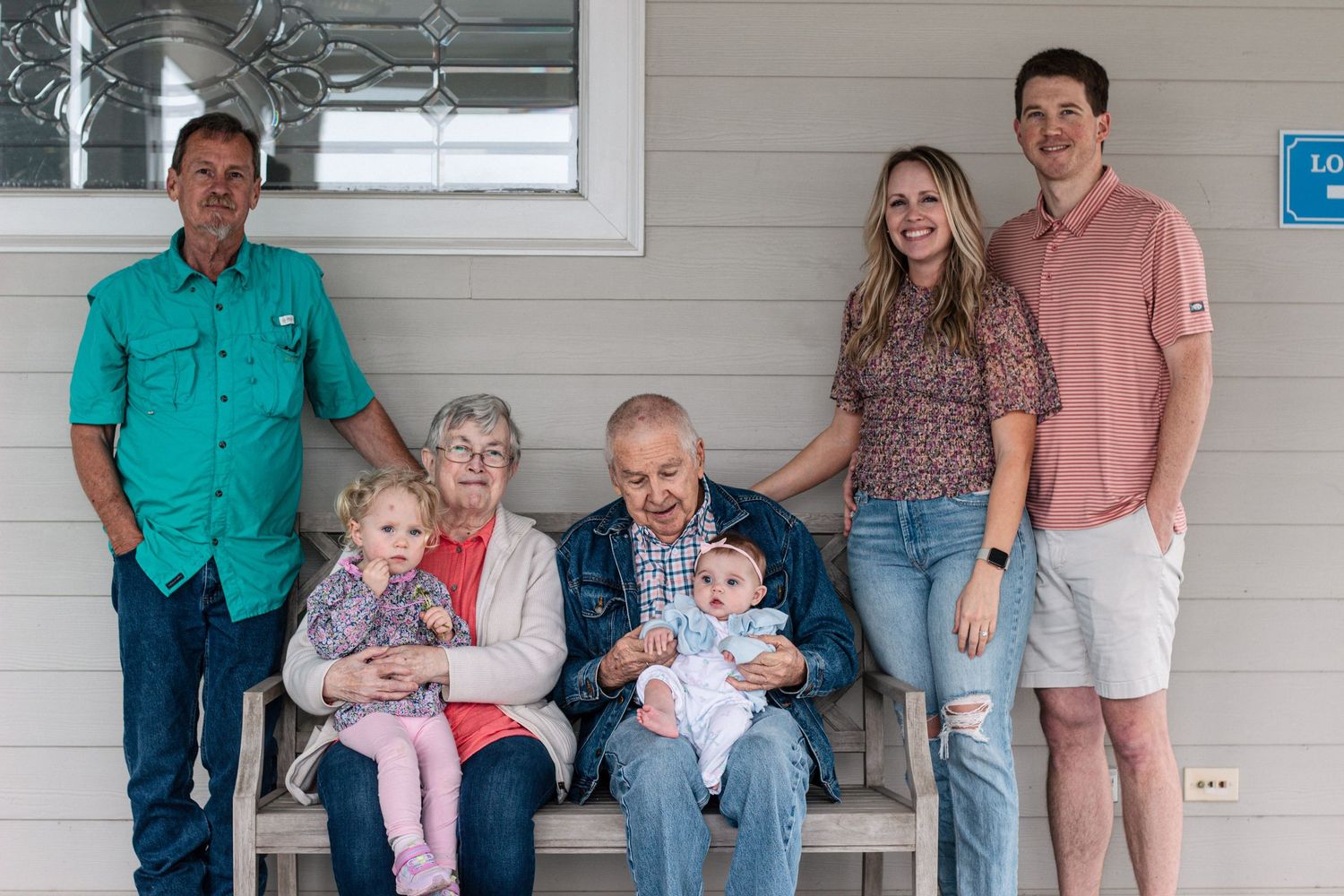 Multi-generational family group portrait on a porch with grandparents holding young children and grandchildren standing nearby.
