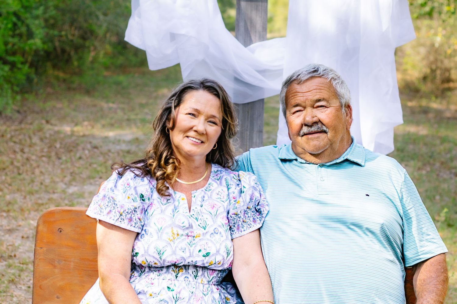 Smiling couple seated on a bench outdoors with white fabric draped behind them in natural setting.