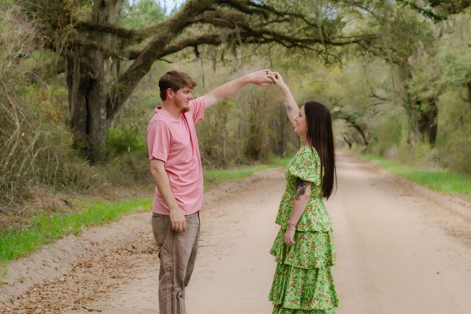 Romantic couple twirling on a tree-lined dirt path, man in pink shirt spinning woman in green tiered floral dress.