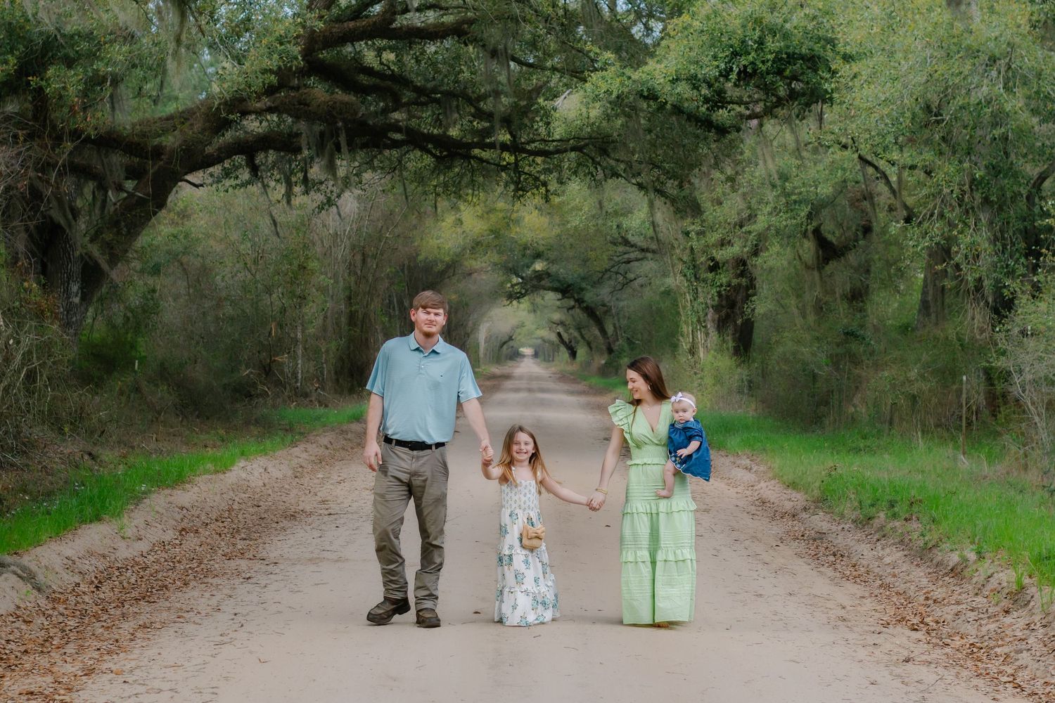 Family of four holding hands on a scenic tree-lined dirt road surrounded by lush green oak trees.