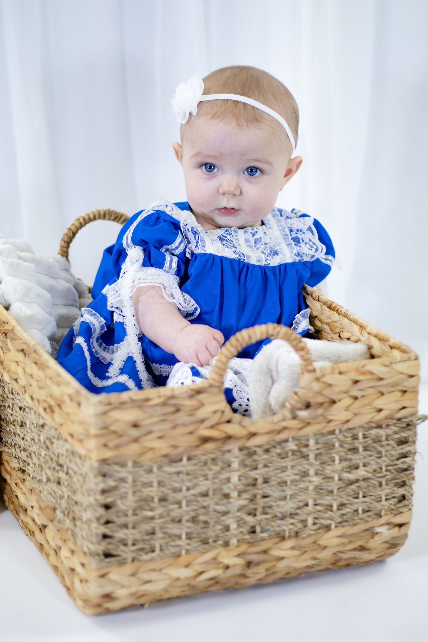 A sweet baby girl in a blue and white lace dress sits in a large rectangular wicker basket.