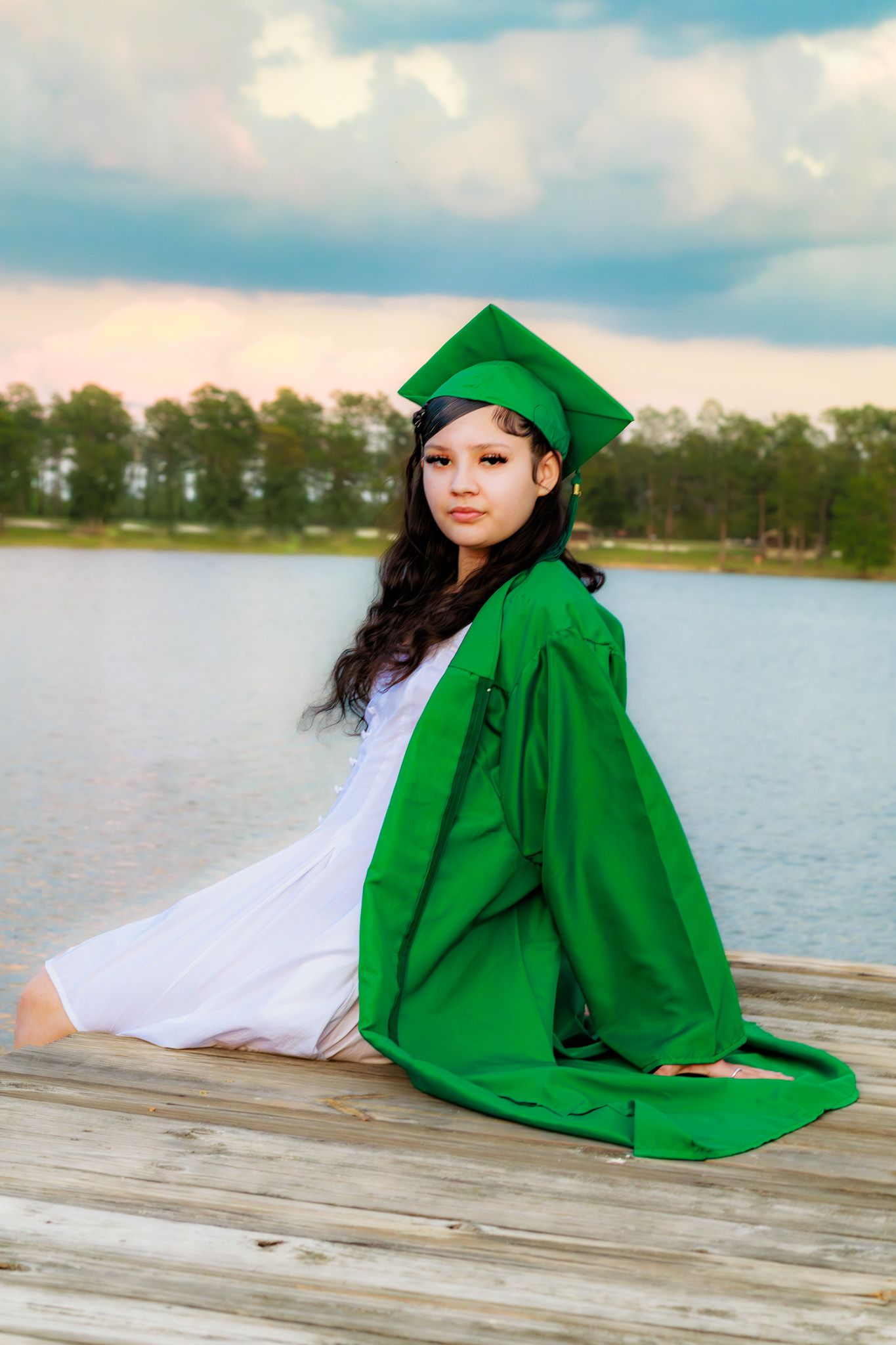 Young girl in green graduation cap and gown sits on wooden dock by lake at sunset.