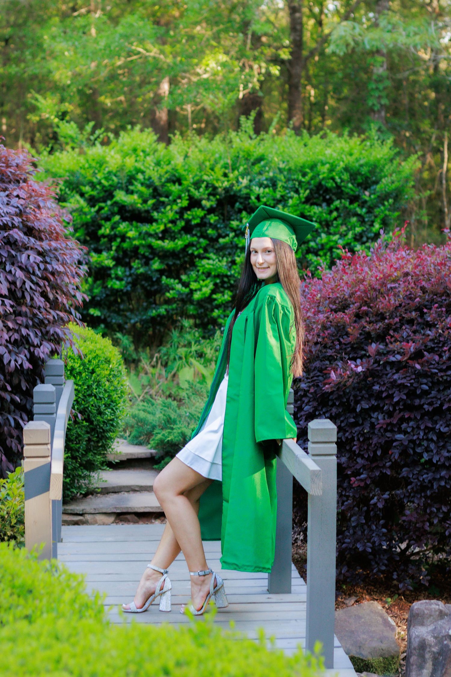 Young woman in green graduation cap and gown poses on decorative wooden bridge surrounded by colorful garden shrubs.