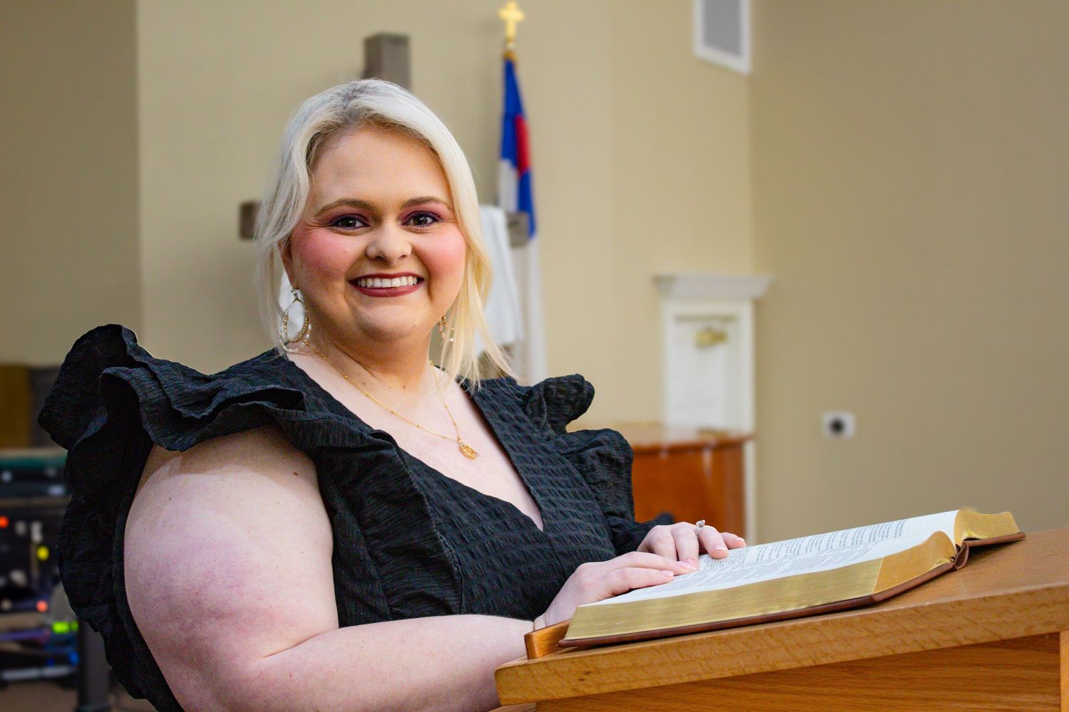 Smiling blonde woman in black ruffled dress posing with open Bible at church lectern.