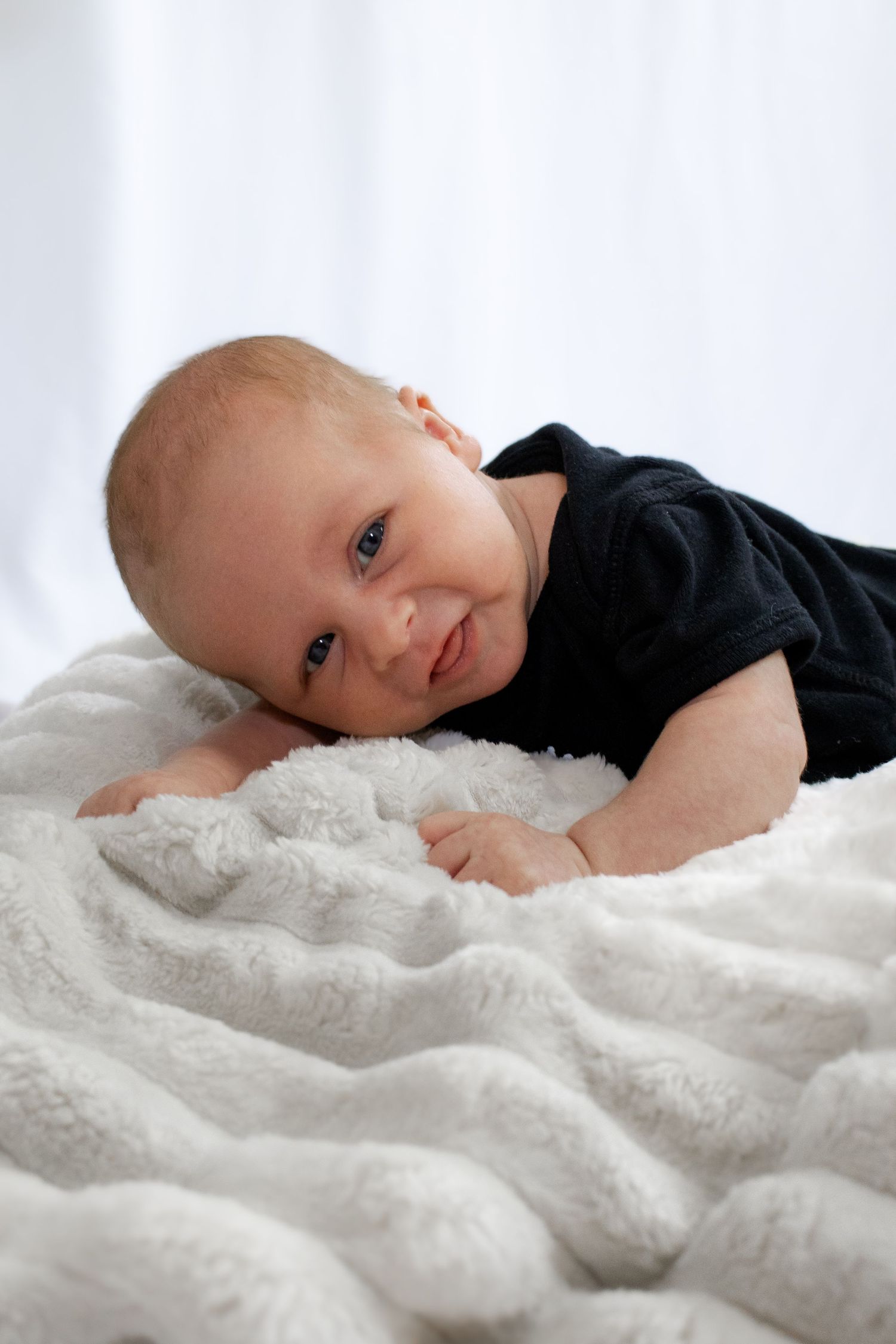 Smiling newborn baby in black onesie lying on a fluffy white blanket.