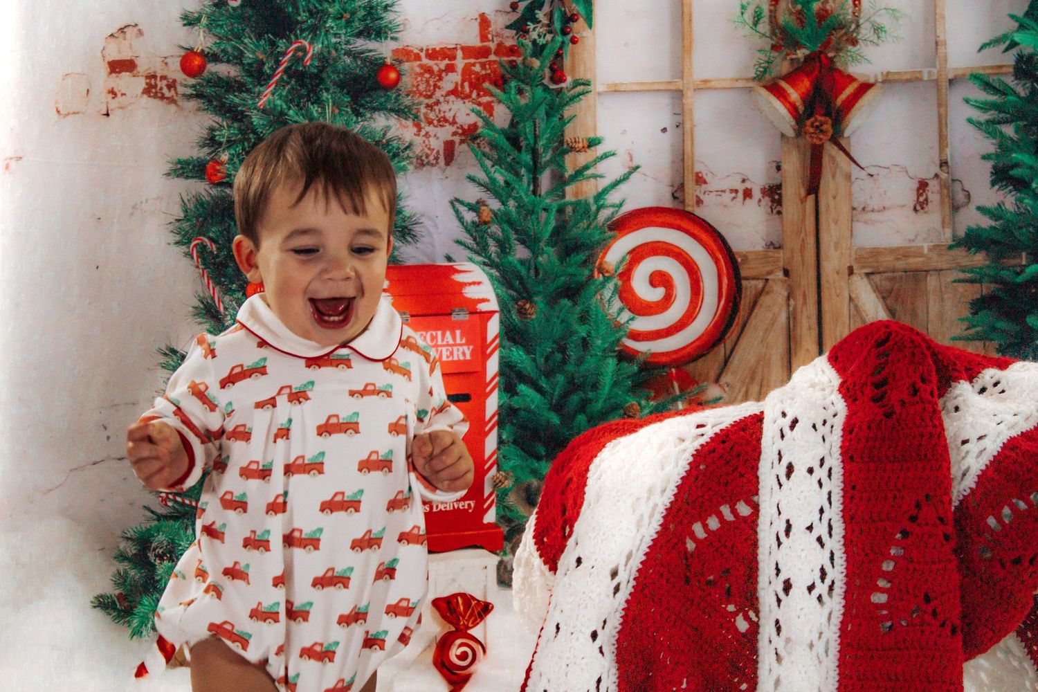 Happy toddler in red truck Christmas pajamas laughing in a festive candy cane holiday photo backdrop.